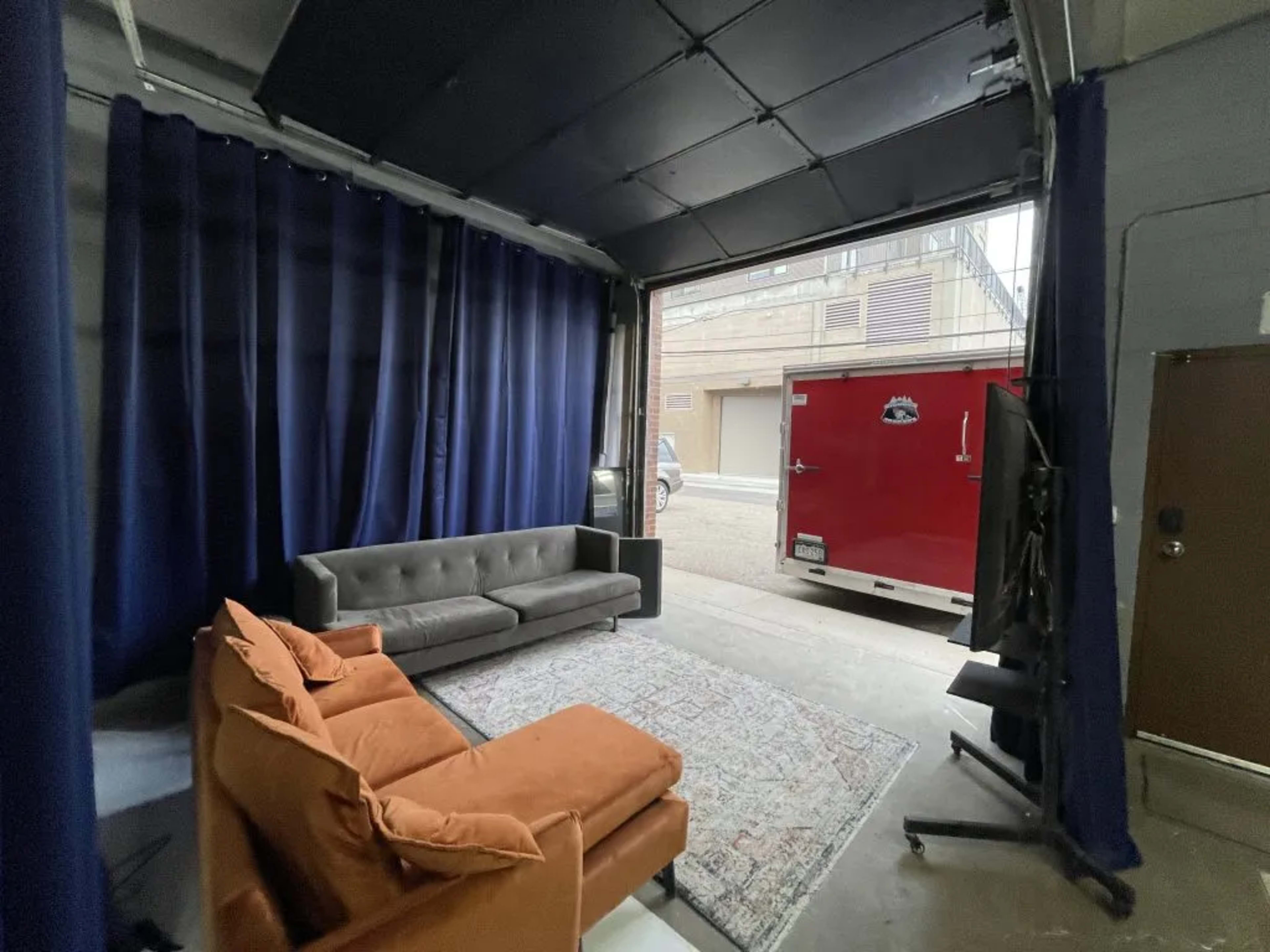 An interior space with a gray couch and an orange armchair, framed by dark curtains and a view of a red trailer outside through an open garage door.