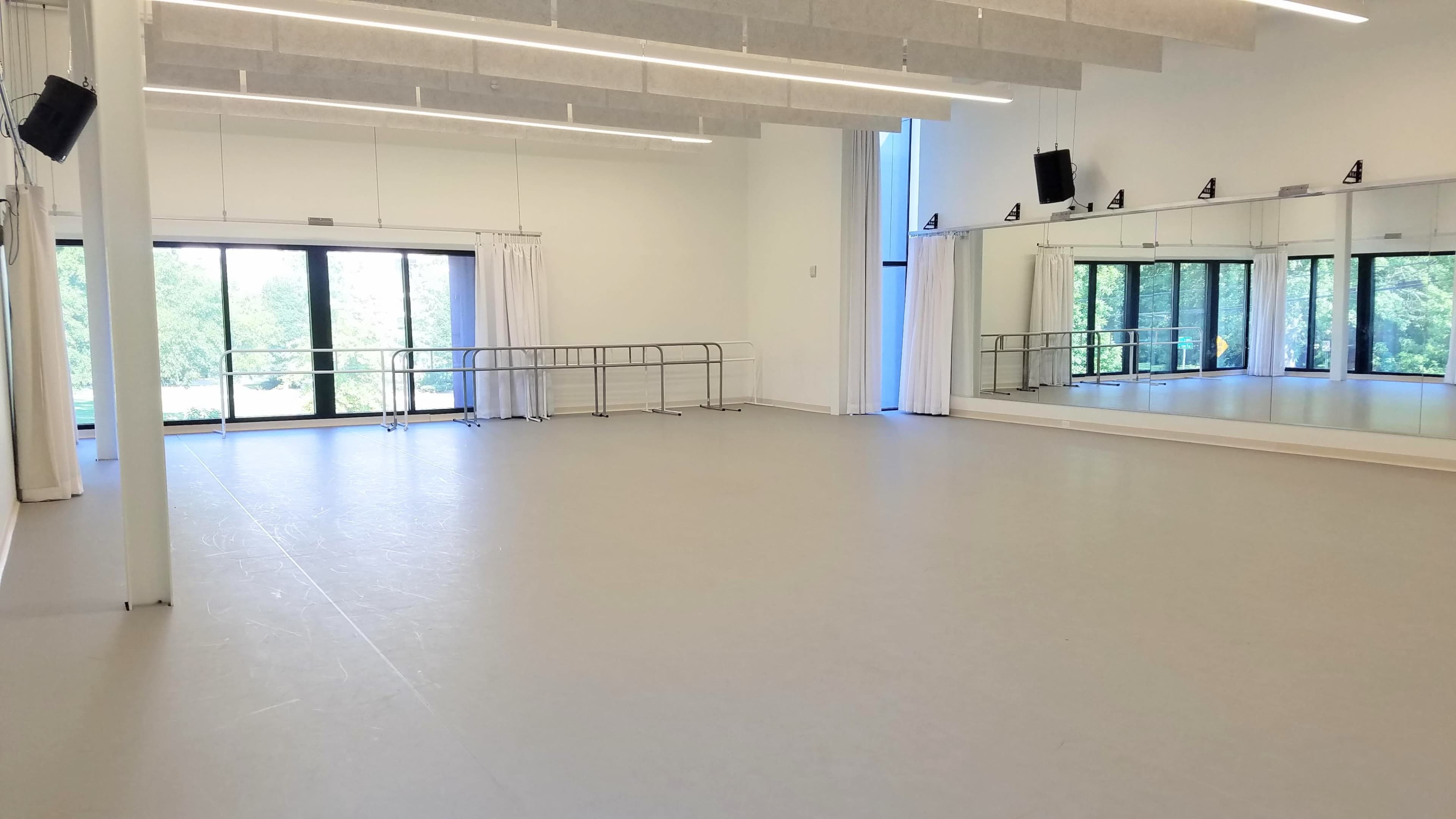 The image shows an empty dance studio with large windows, mirrors on one wall, and a barre along the floor.