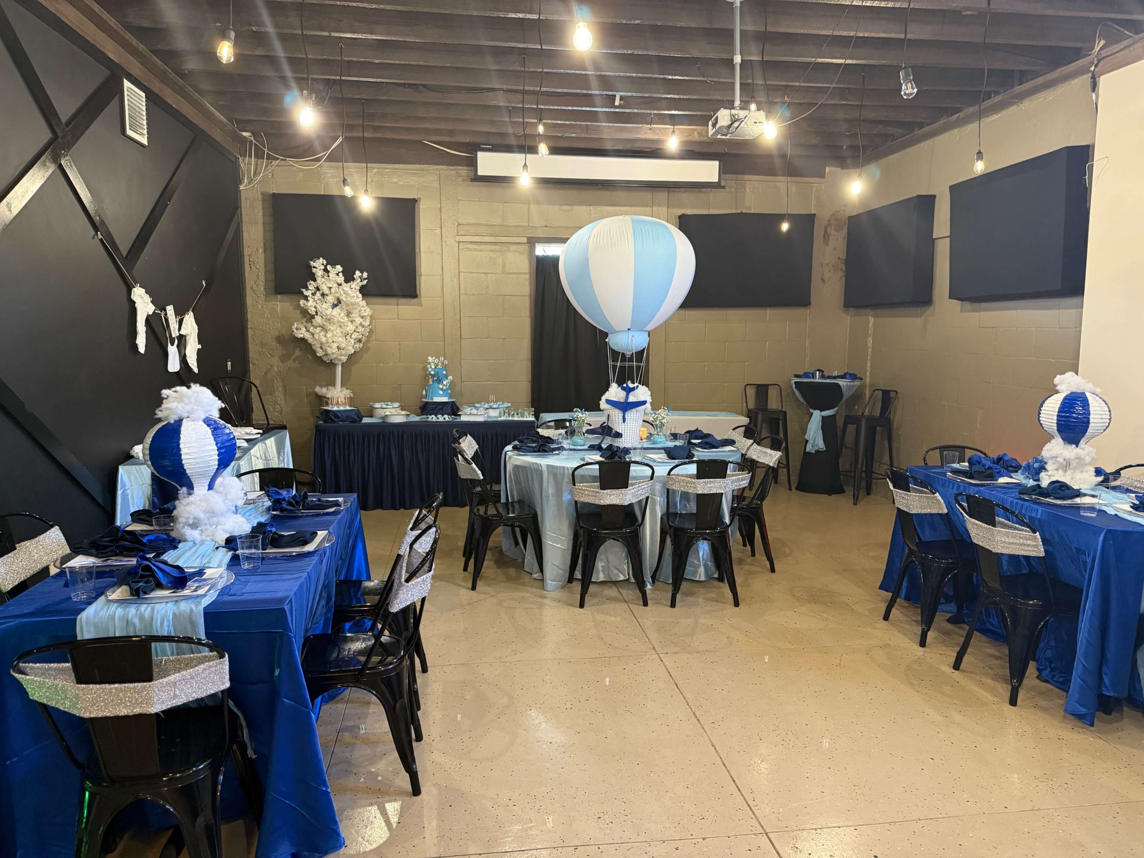 The image shows a decorated indoor event space featuring tables with blue and white tablecloths, black chairs, and large balloon centerpieces.