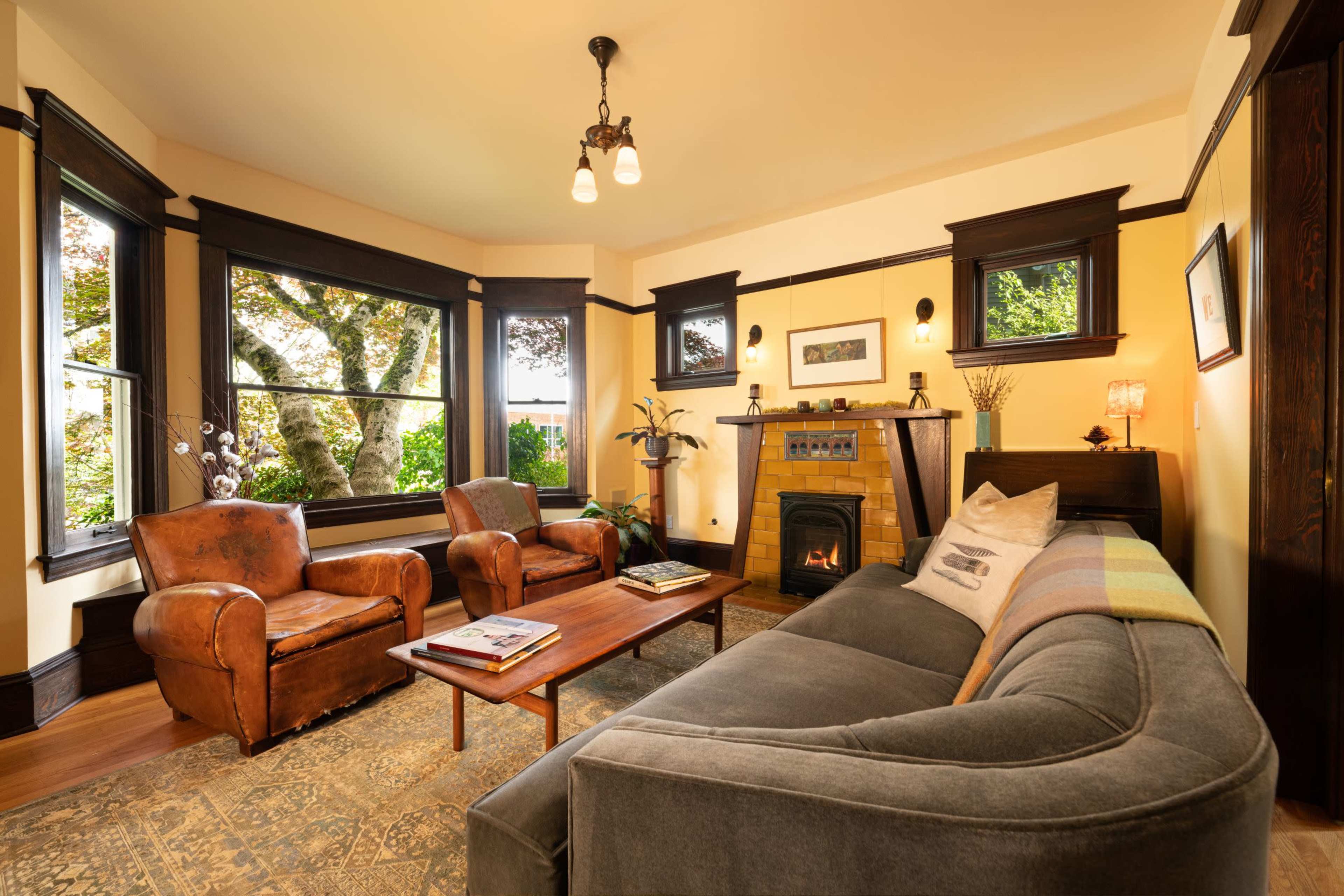 A cozy living room with two armchairs and a sofa arranged around a central coffee table, featuring a fireplace and windows that provide a view of the outdoors.