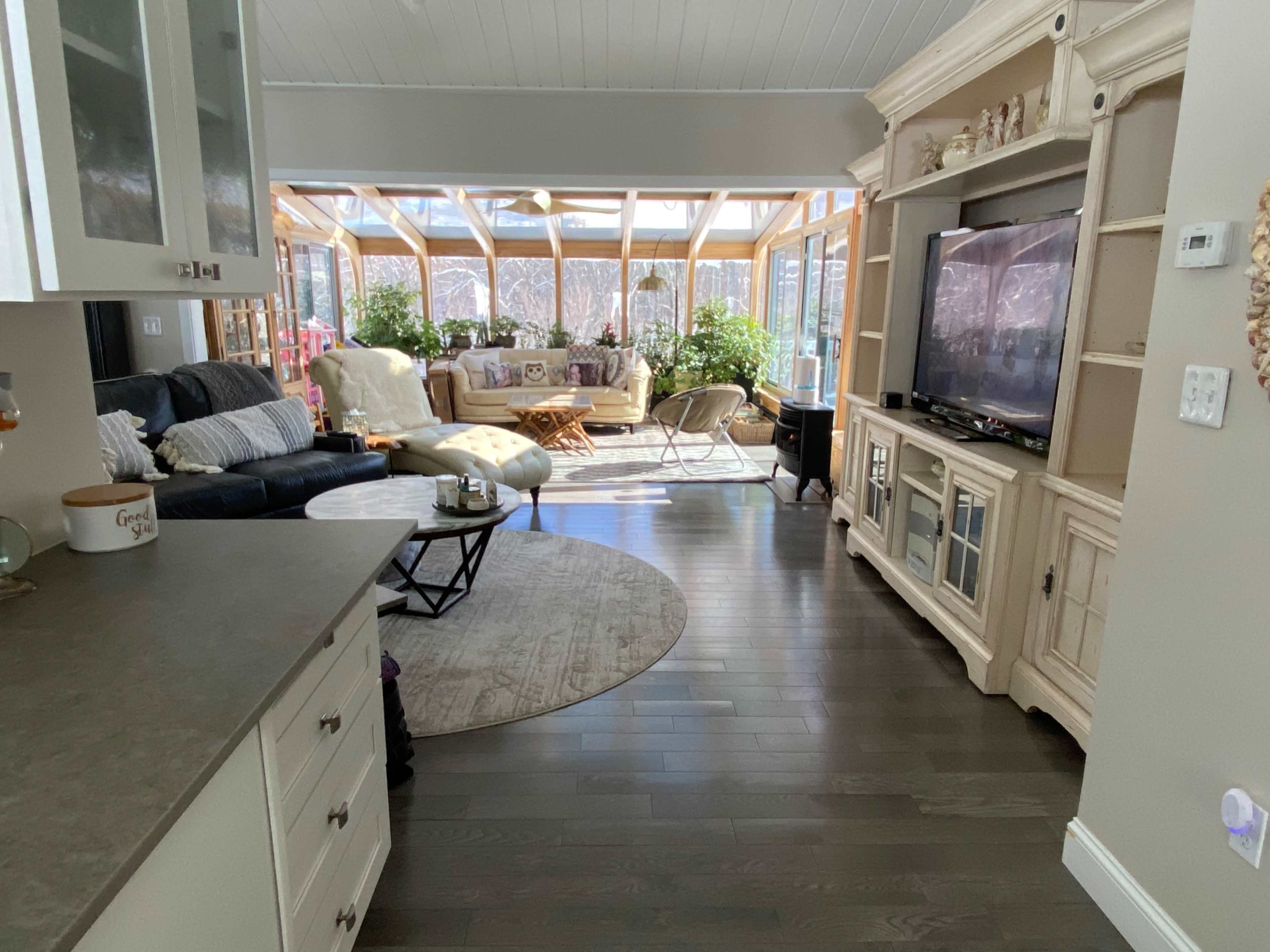 A spacious living area with light-colored furniture, a large television, and a sunroom filled with plants and natural light.