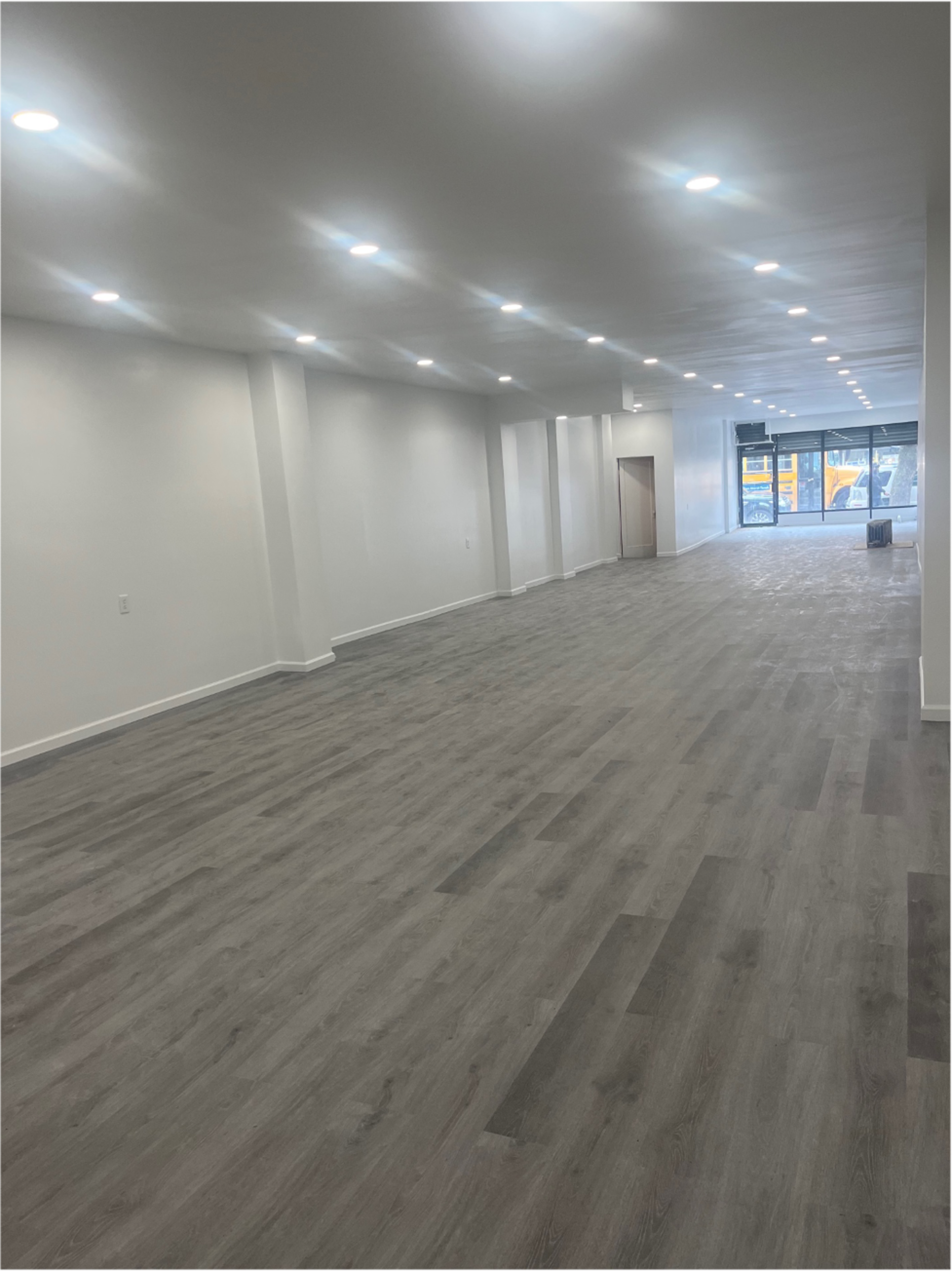 A spacious, empty interior with light-colored walls, a wooden floor, and rows of ceiling lights illuminating the area.