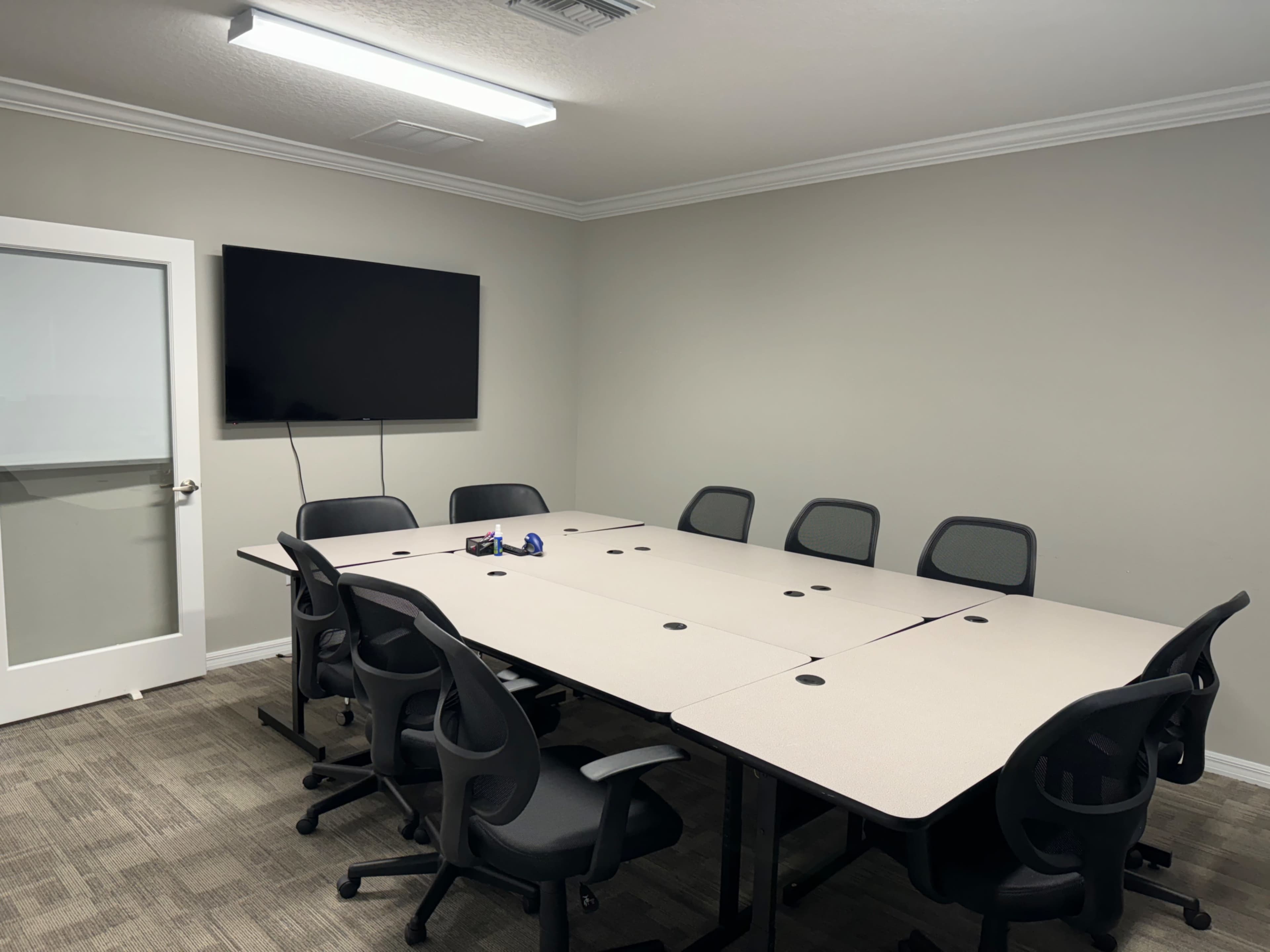 A conference room features a large table surrounded by ergonomic chairs, with a TV mounted on the wall and a glass door in the background.