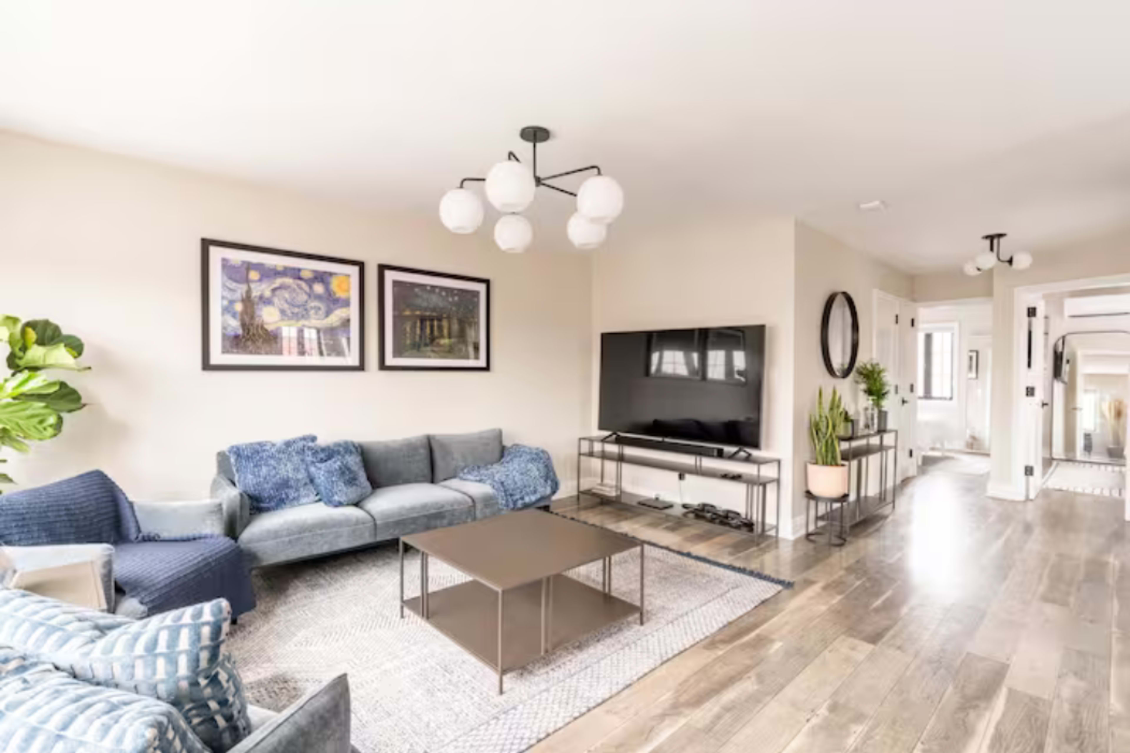 A modern living room features a gray sectional sofa, a coffee table, and a television on a stand, with artwork on the walls and a plant in the corner.