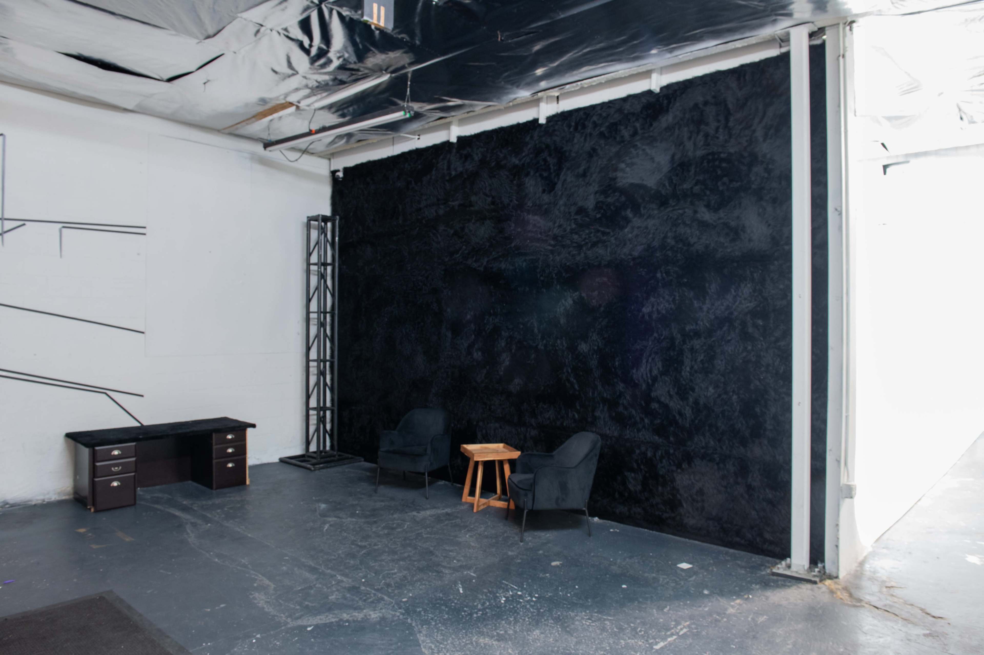 A minimalist studio space with a black wall, two chairs, a small table, and a desk on one side.