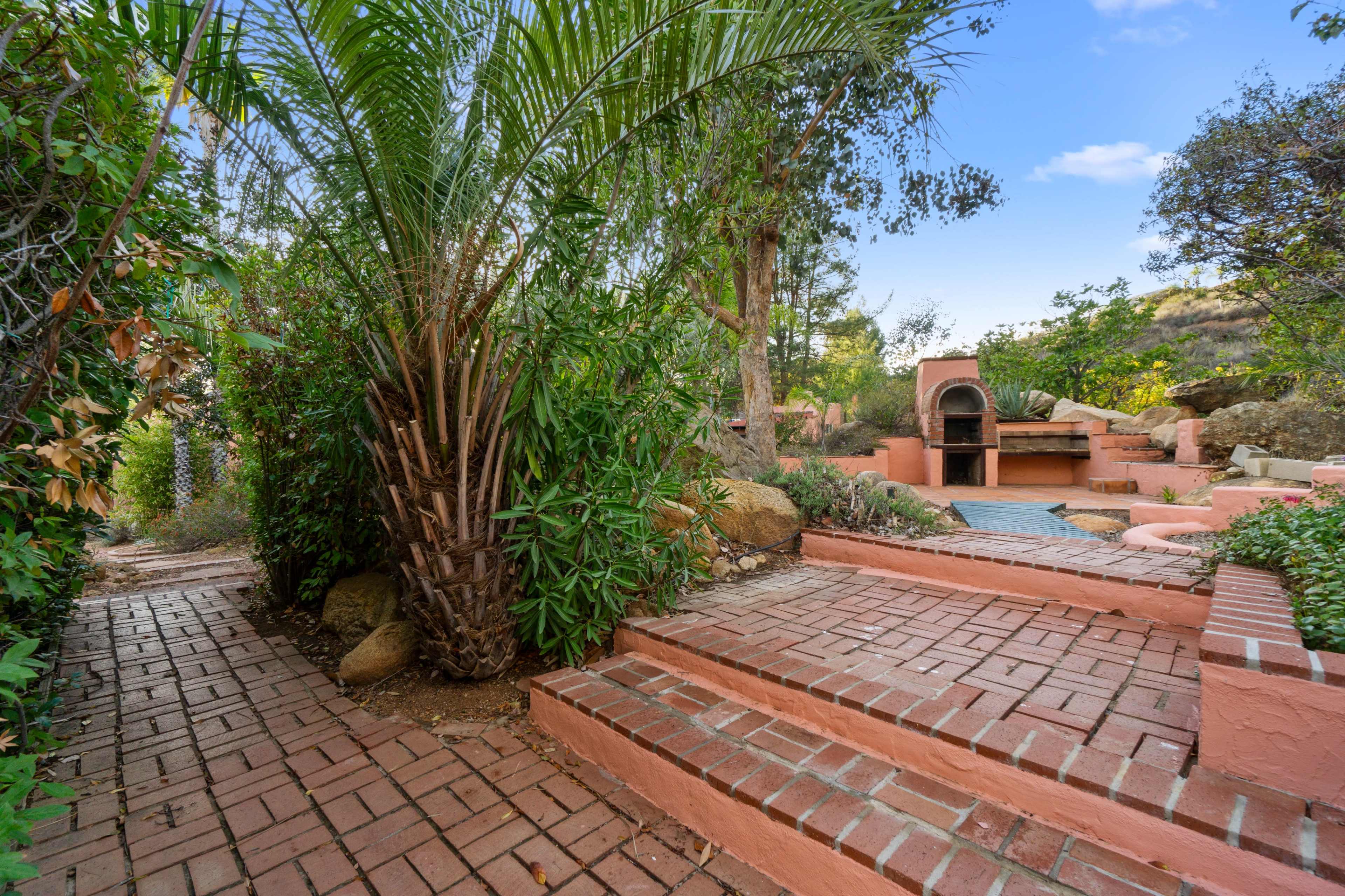 A brick pathway leads through lush greenery to a stone barbecue area amidst natural landscaping.