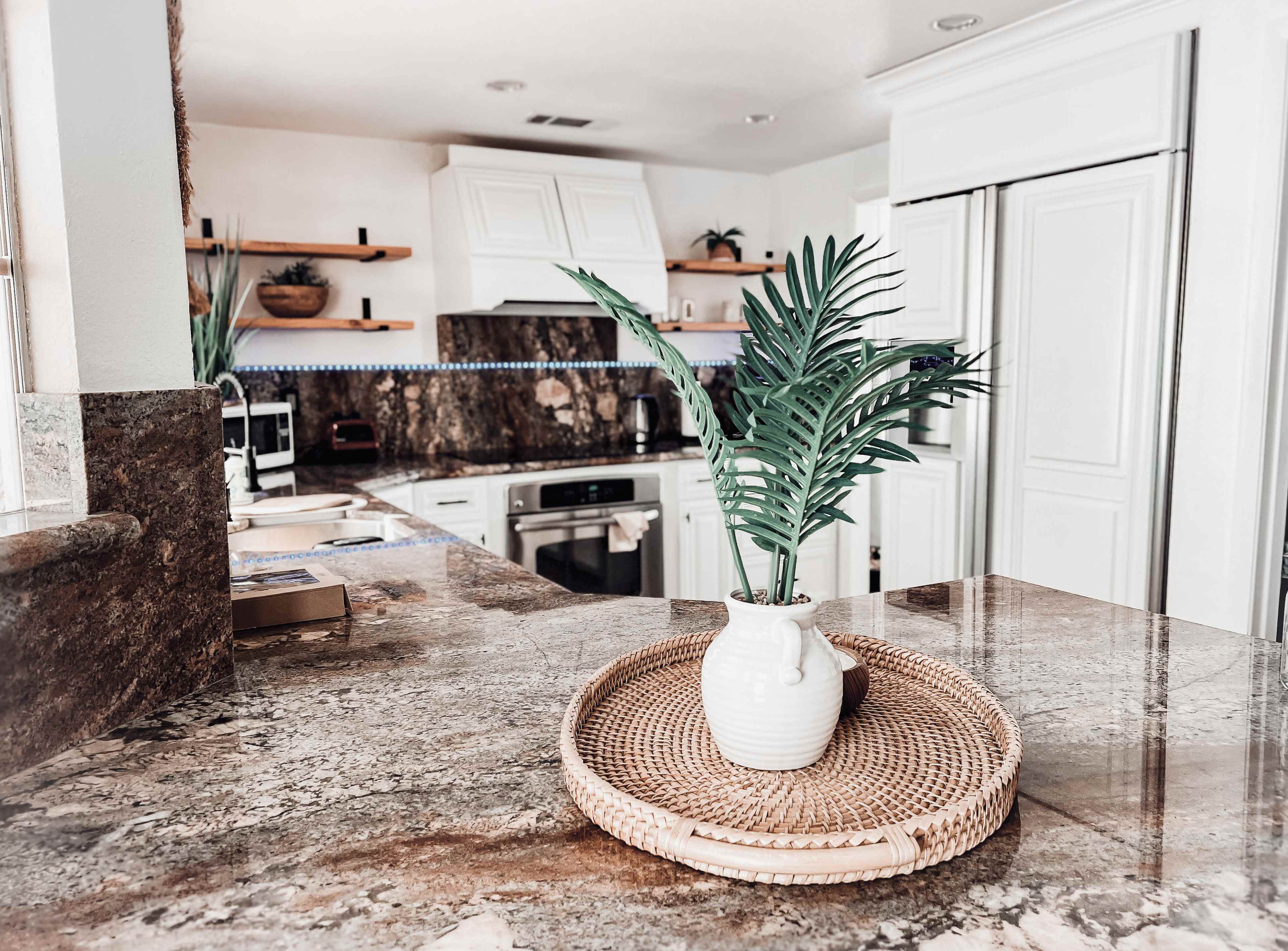 A kitchen features a marble countertop with a woven tray holding a small plant in a white vase, complemented by white cabinetry and shelves.
