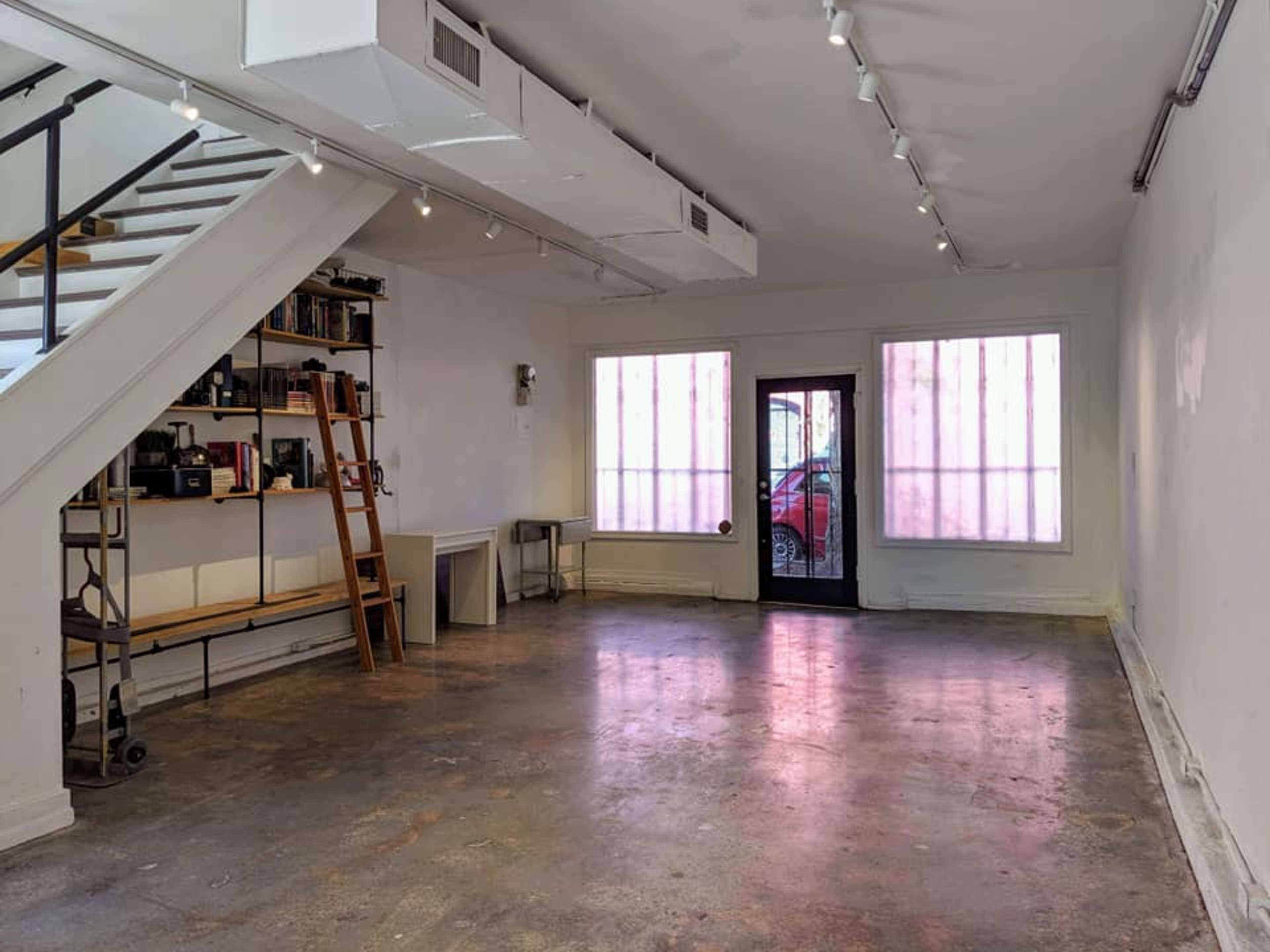 The image shows an empty room with concrete flooring, a staircase on the left, shelves filled with items, and large windows in the front facing a parked car outside.