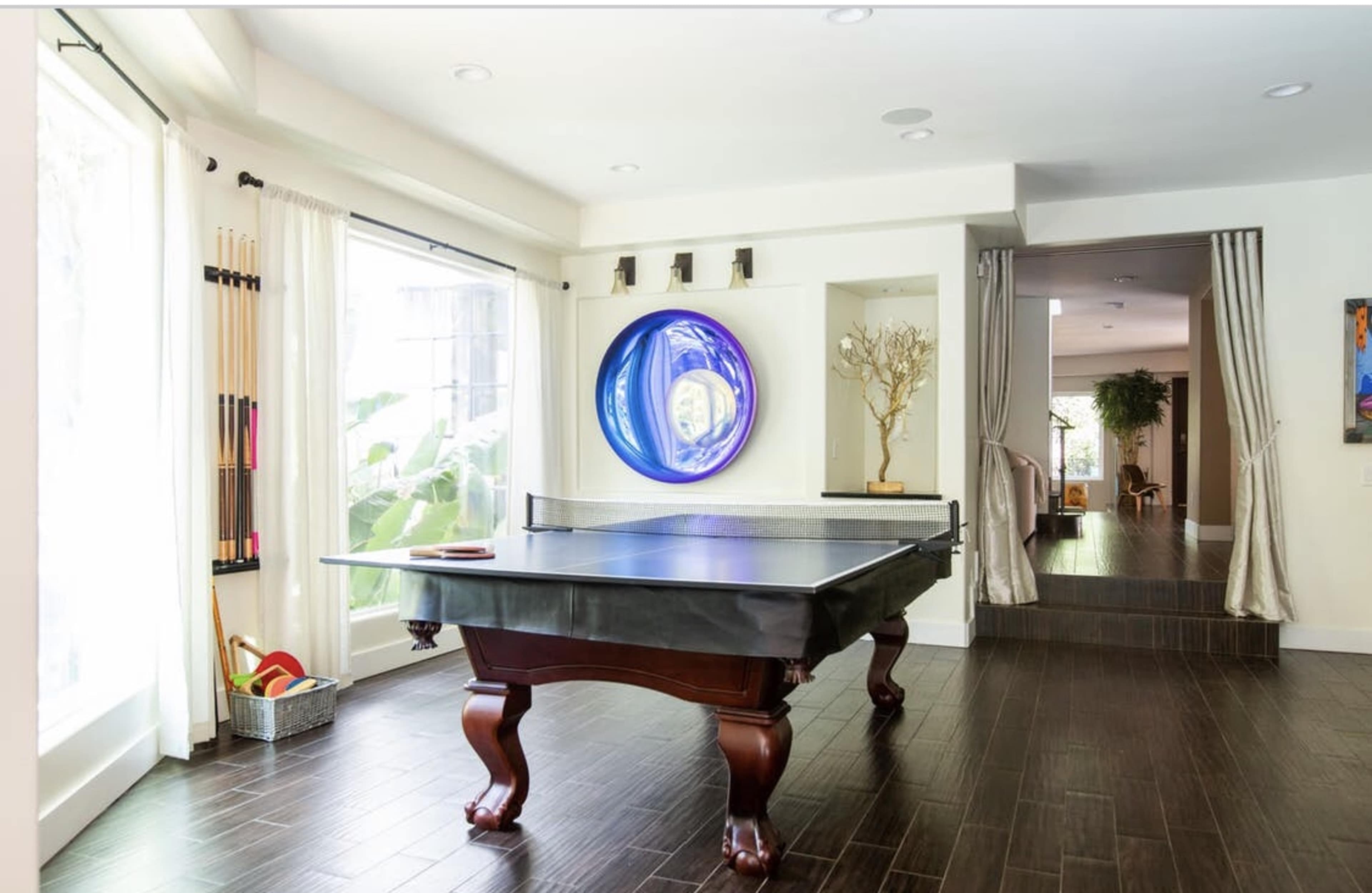 A room featuring a pool table surrounded by large windows and modern decor, with a hallway leading to another area visible in the background.