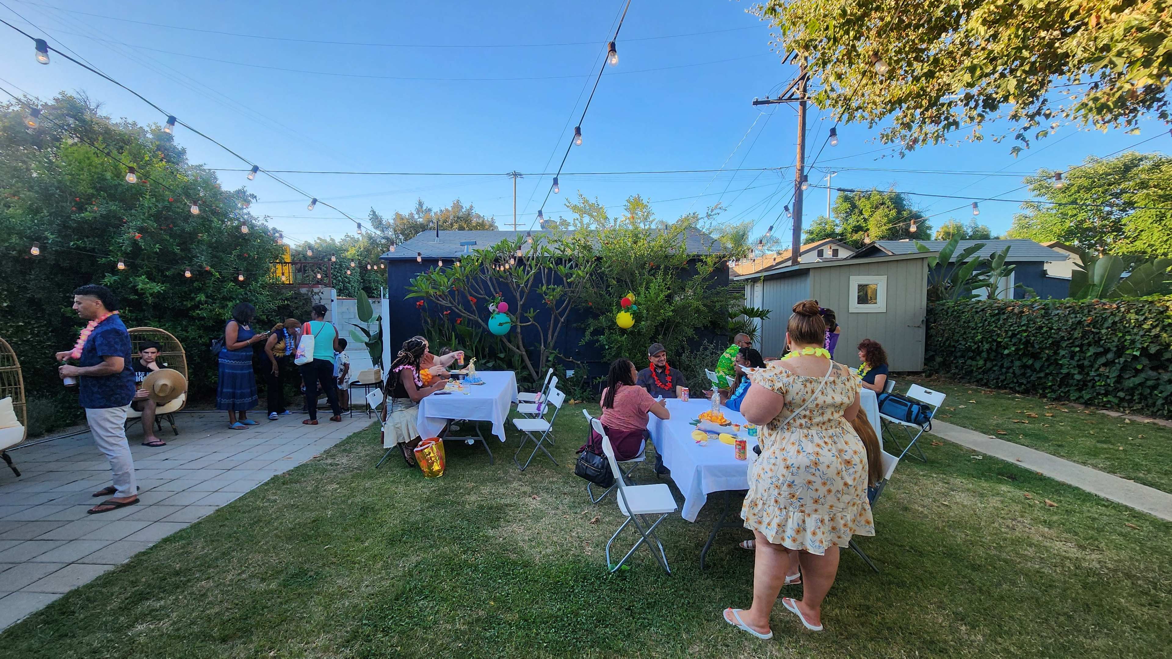 A group of people gathers in a backyard decorated with string lights, enjoying a festive outdoor event with tables set for dining.