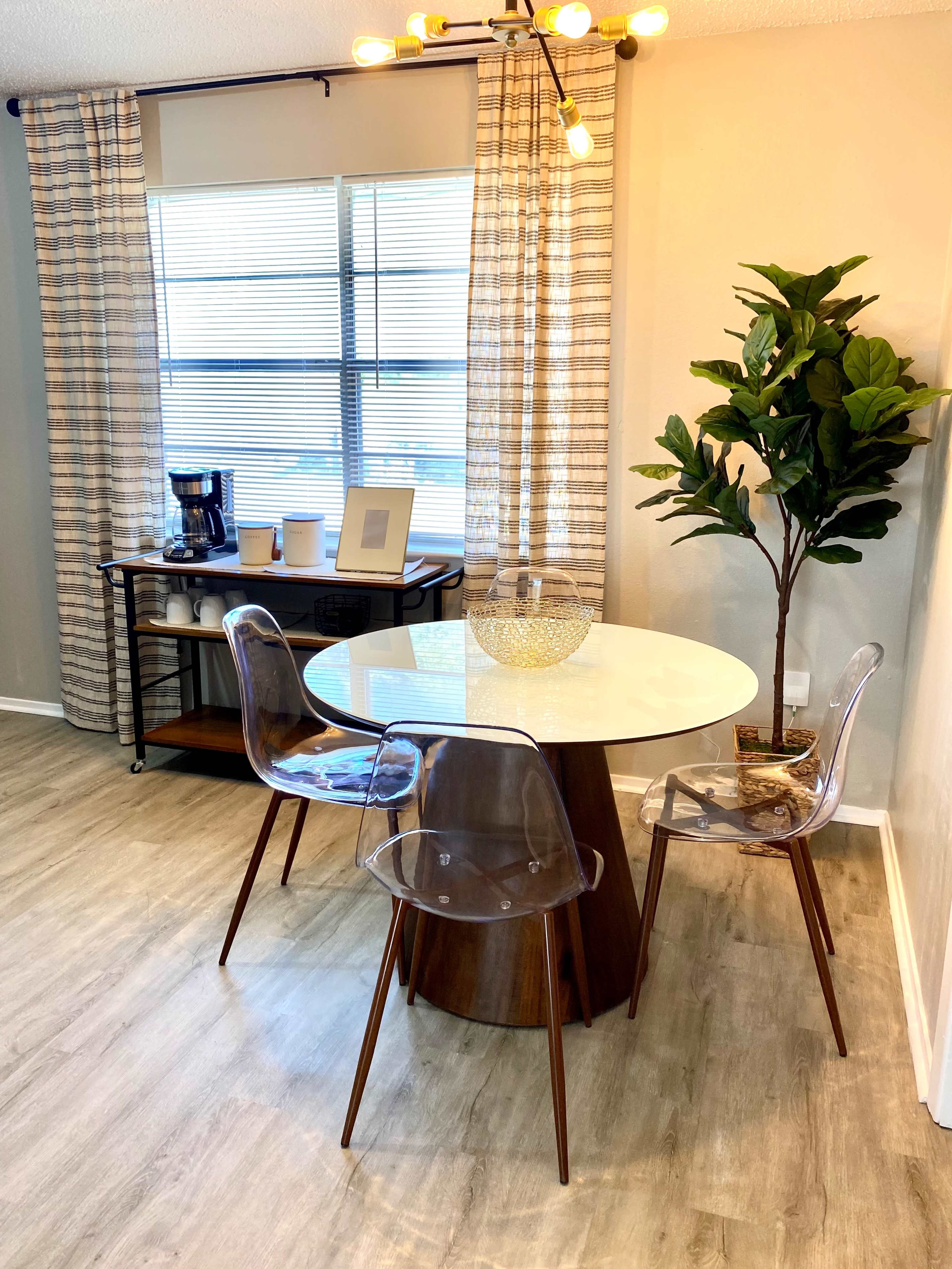 A round white table with three clear chairs is positioned near a window with sheer curtains, accompanied by a coffee station and a potted plant.