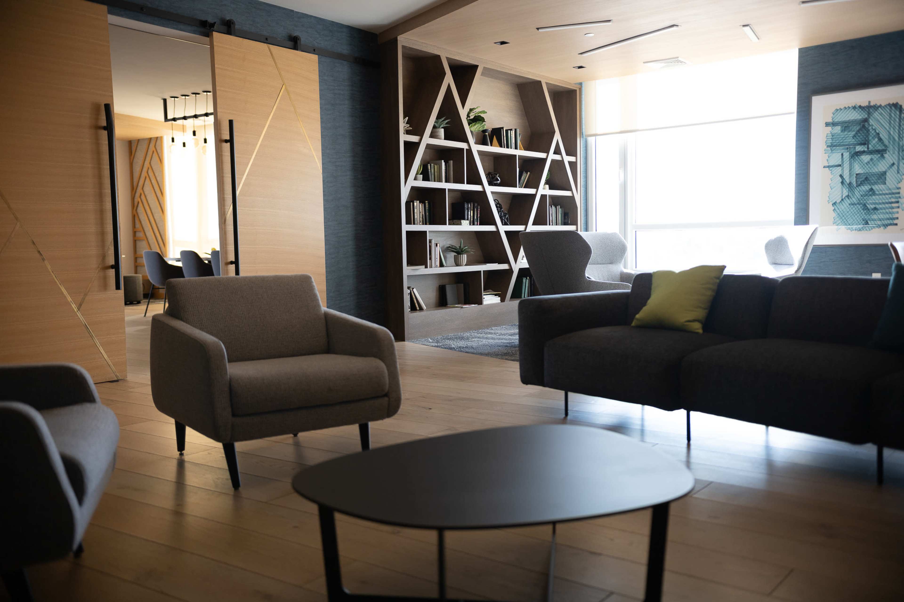 The image shows a modern, open-concept lounge area with a coffee table, two armchairs, and a large bookshelf, alongside a view of a dining space in the background.