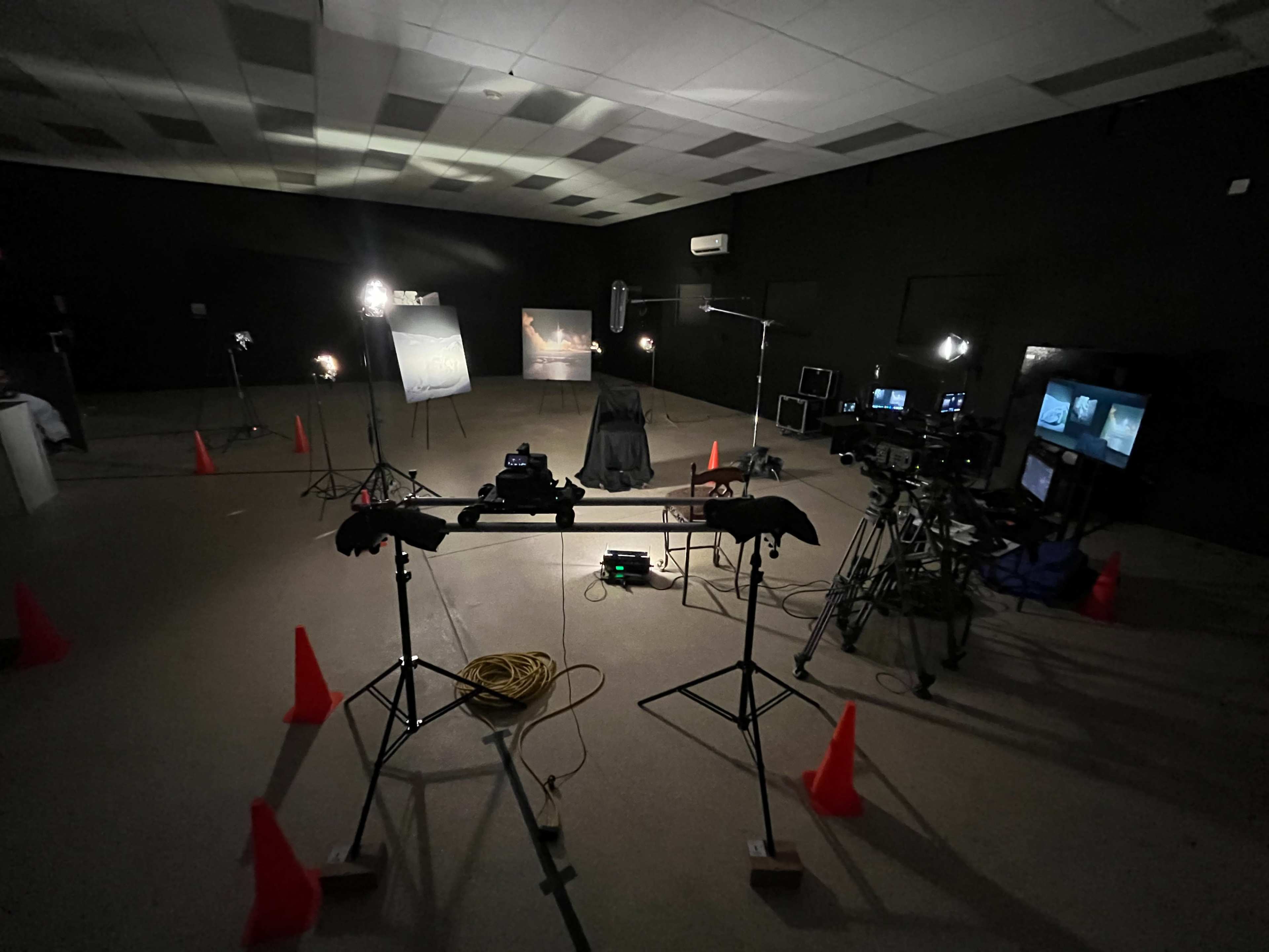 A dimly lit studio setup with cameras, lighting equipment, and orange cones arranged around a central chair and backdrop.