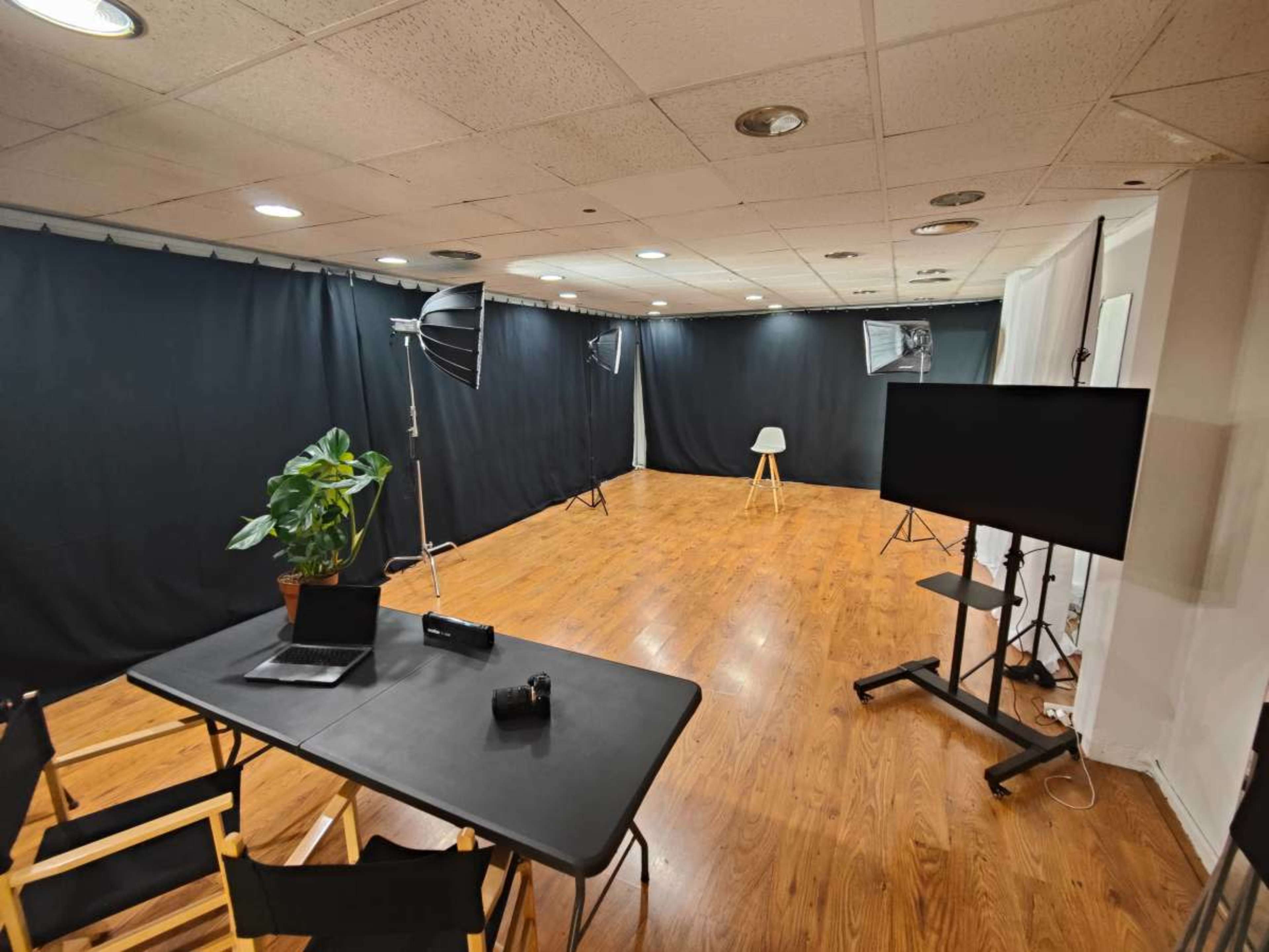 The image shows a spacious, well-lit room with black curtains, professional lighting equipment, a table with a laptop and camera, and a chair positioned in the center.