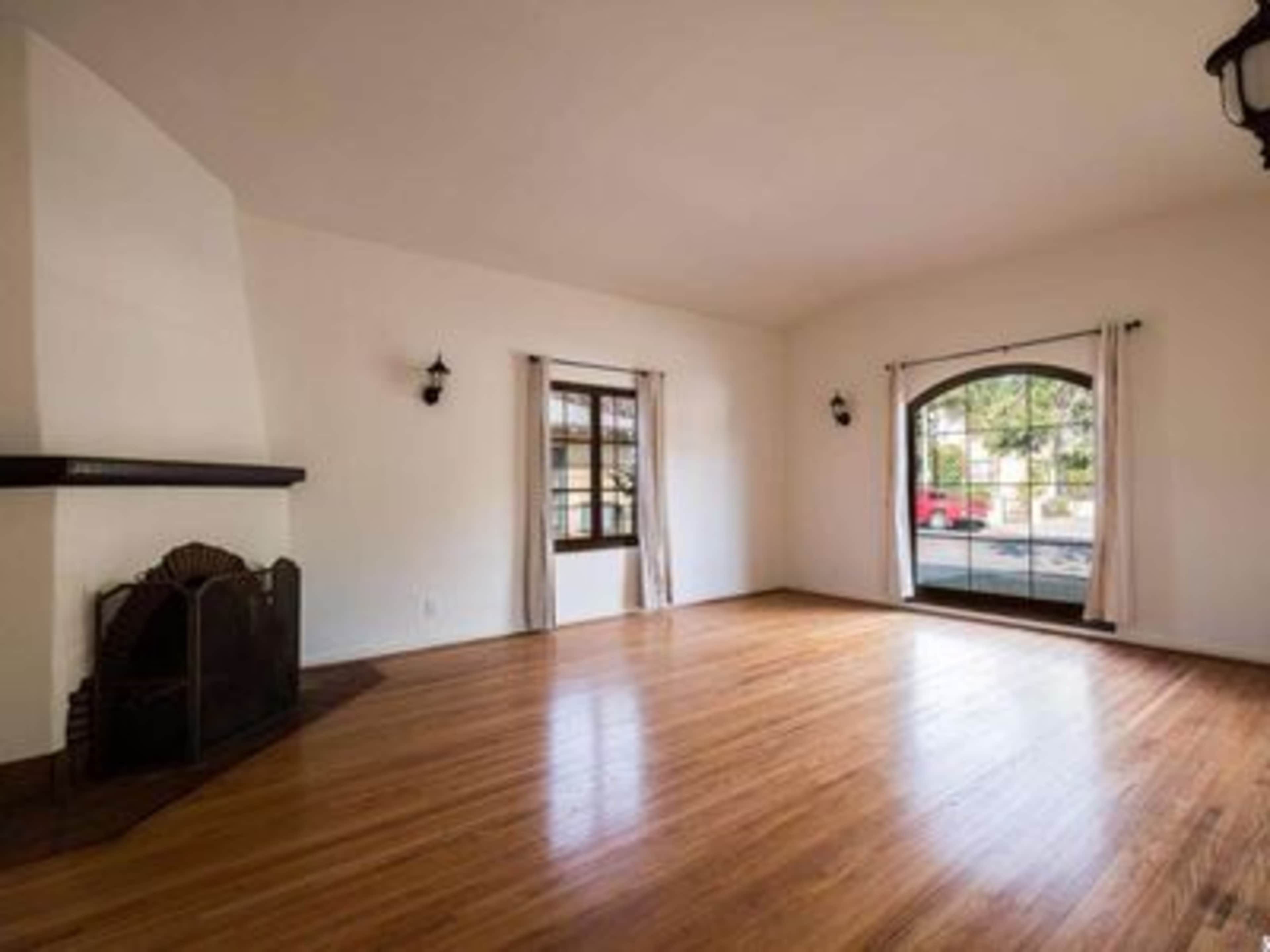 An empty living room with wooden floors, a fireplace on one side, and a large arched window allowing natural light to enter.