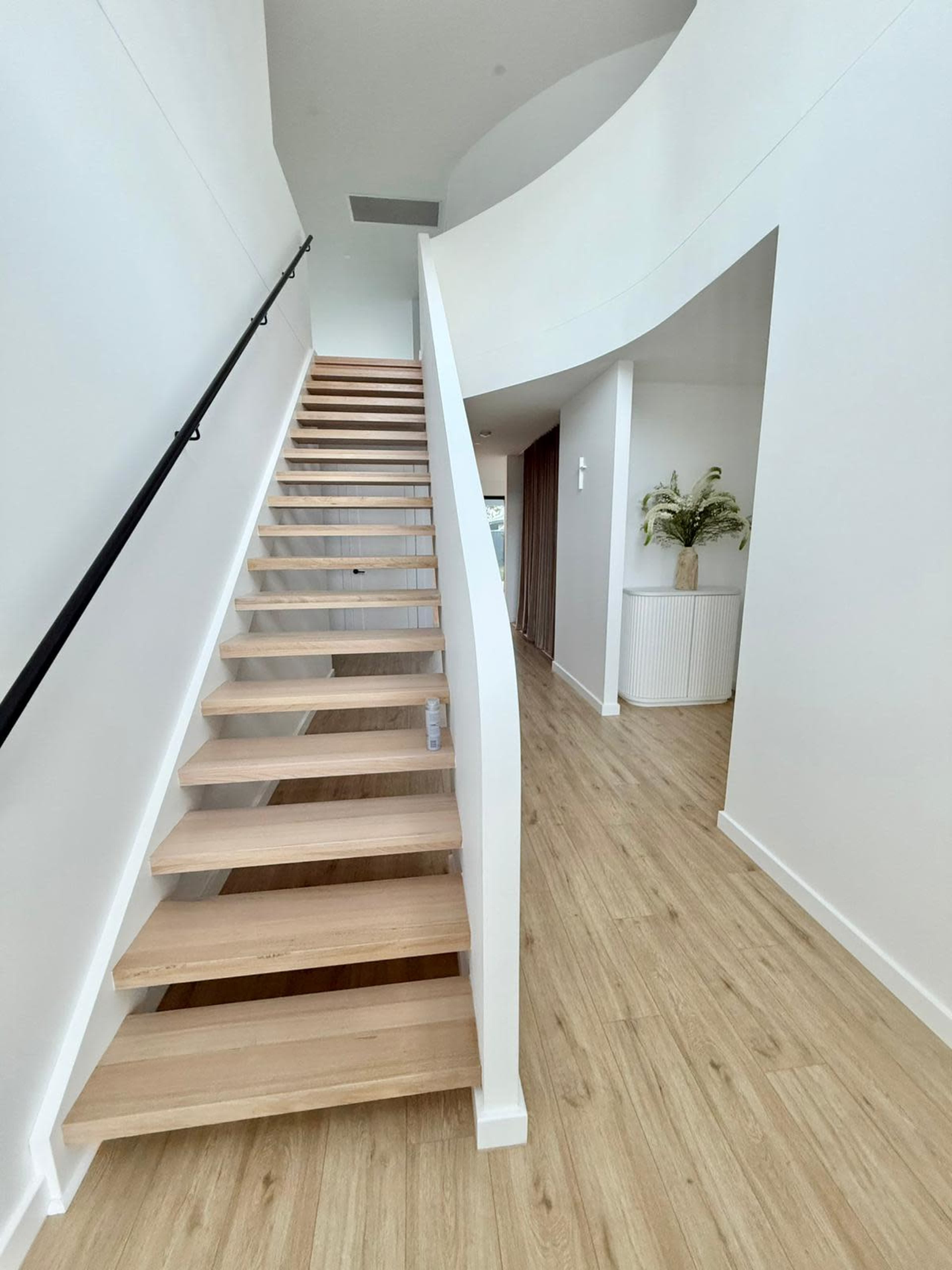 A modern, open staircase with wooden steps leads up to a bright, spacious area accented by smooth white walls and wooden flooring.