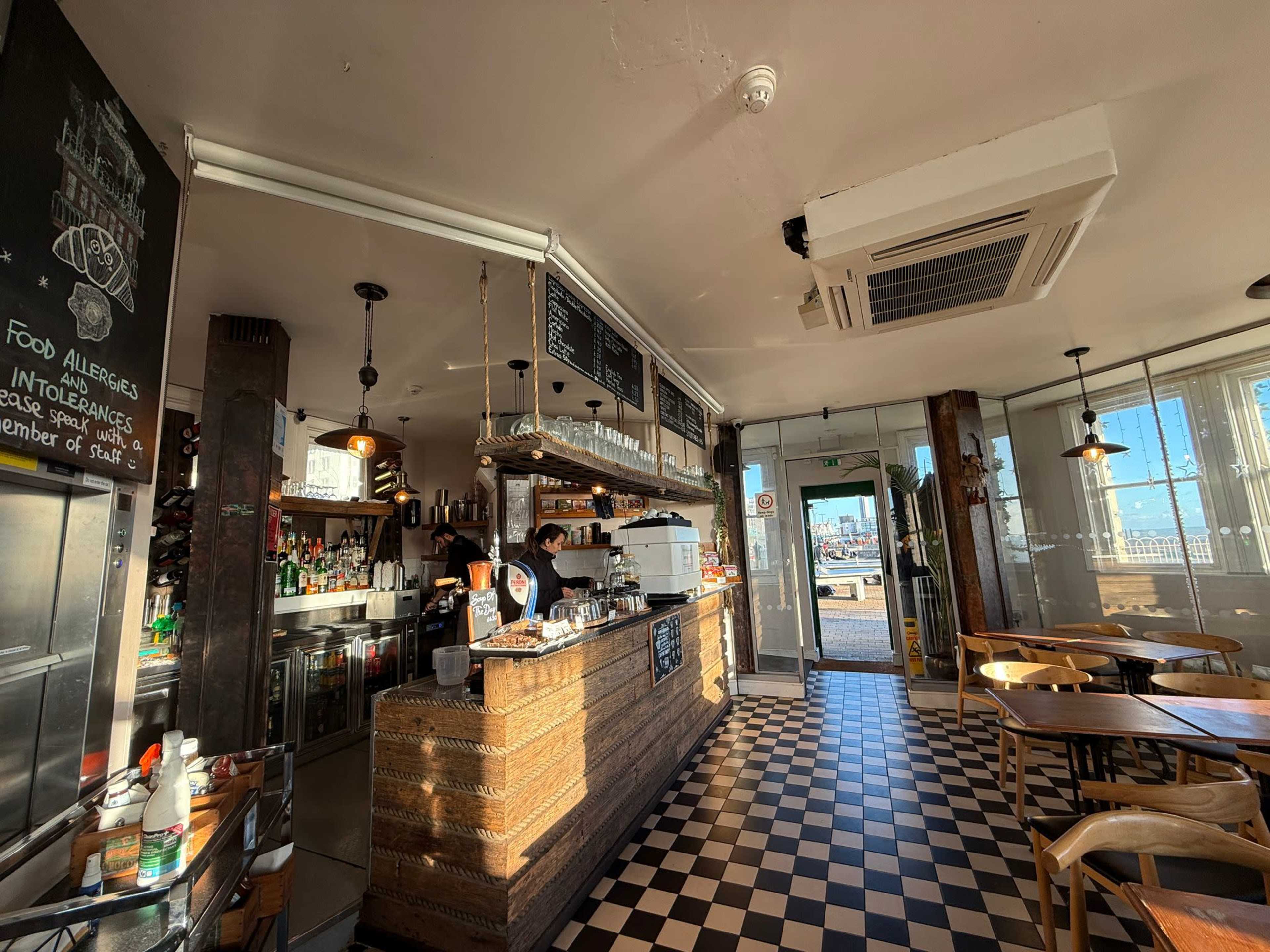 The image shows a cozy café interior with a wooden counter, shelves of drinks, and a checkered floor, while a barista prepares orders behind the counter.
