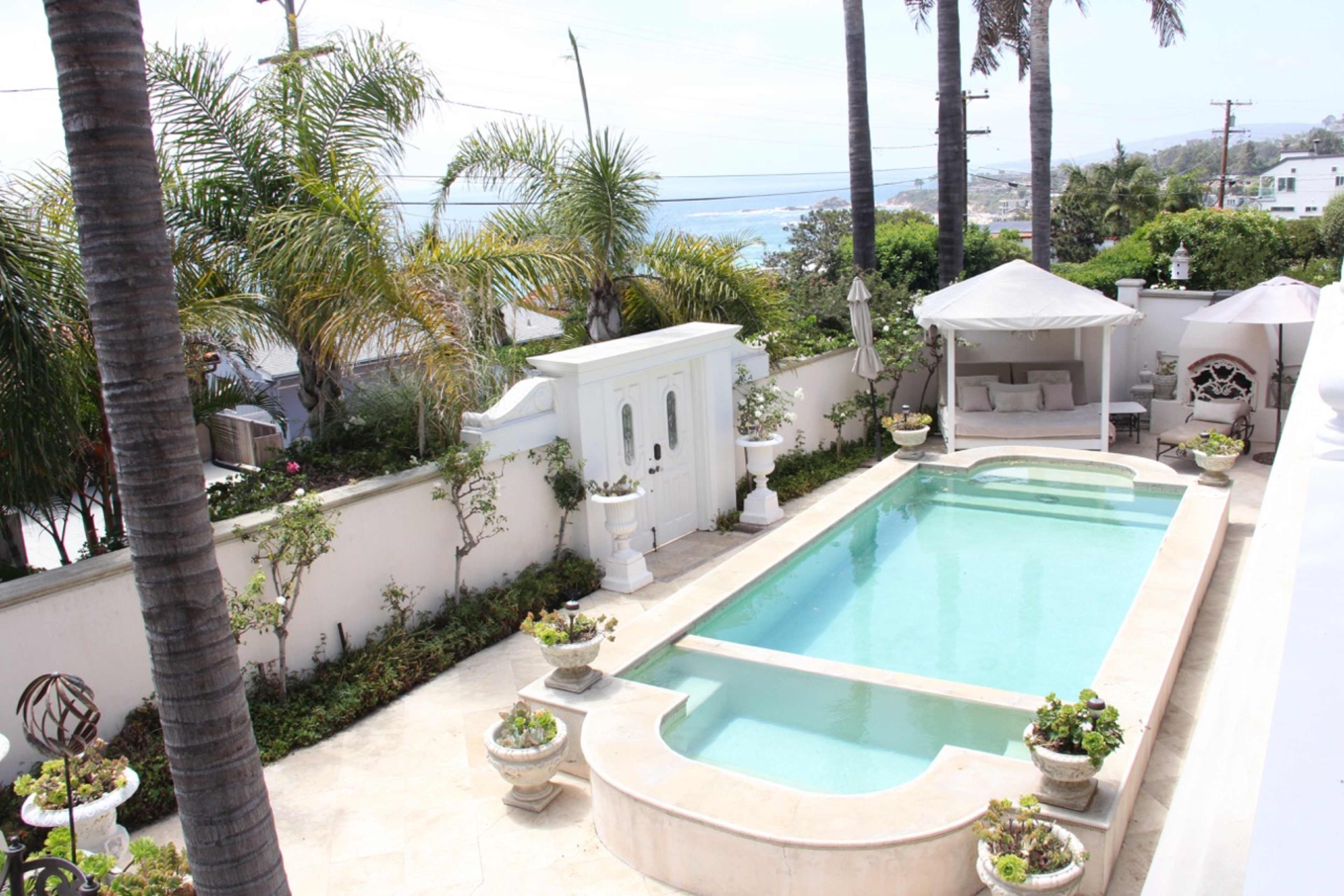 A residential backyard with a swimming pool surrounded by manicured plants and palm trees, overlooking a coastal view.