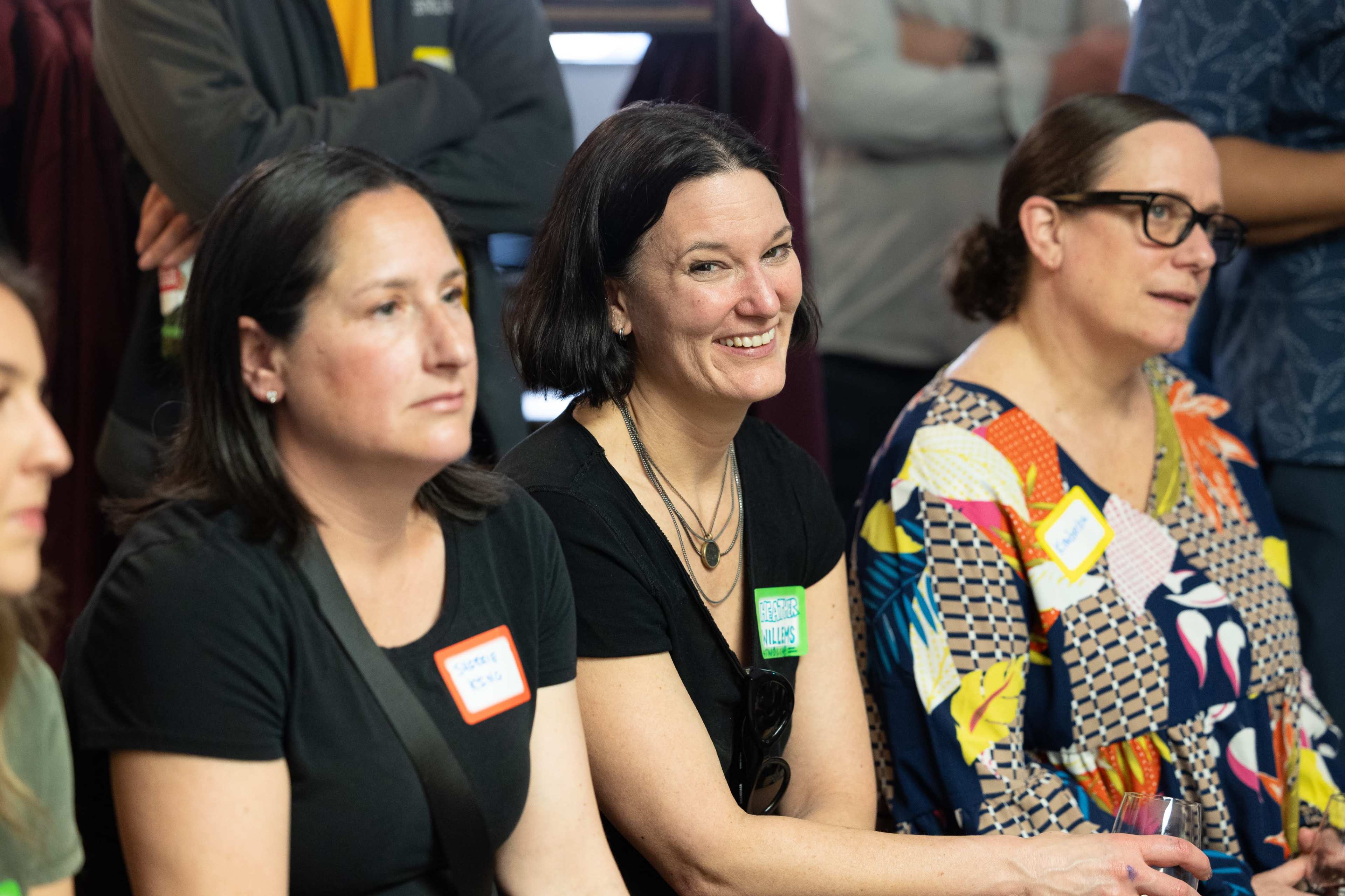 A group of women sit together, smiling and engaging in conversation at a gathering, with name tags visible on their clothing.