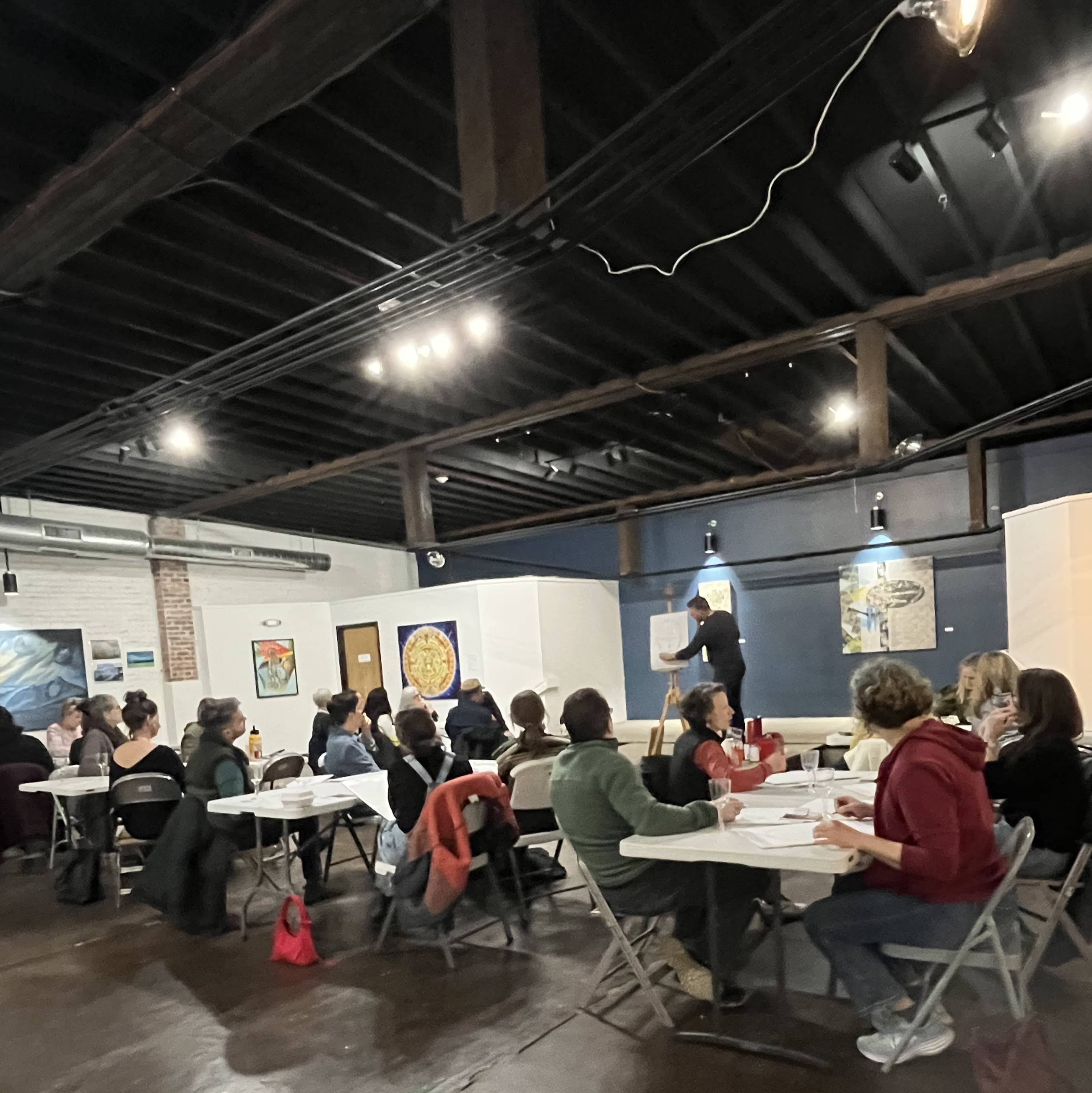 A group of people is seated at tables in a well-lit room, watching a speaker who is presenting on a flip chart.