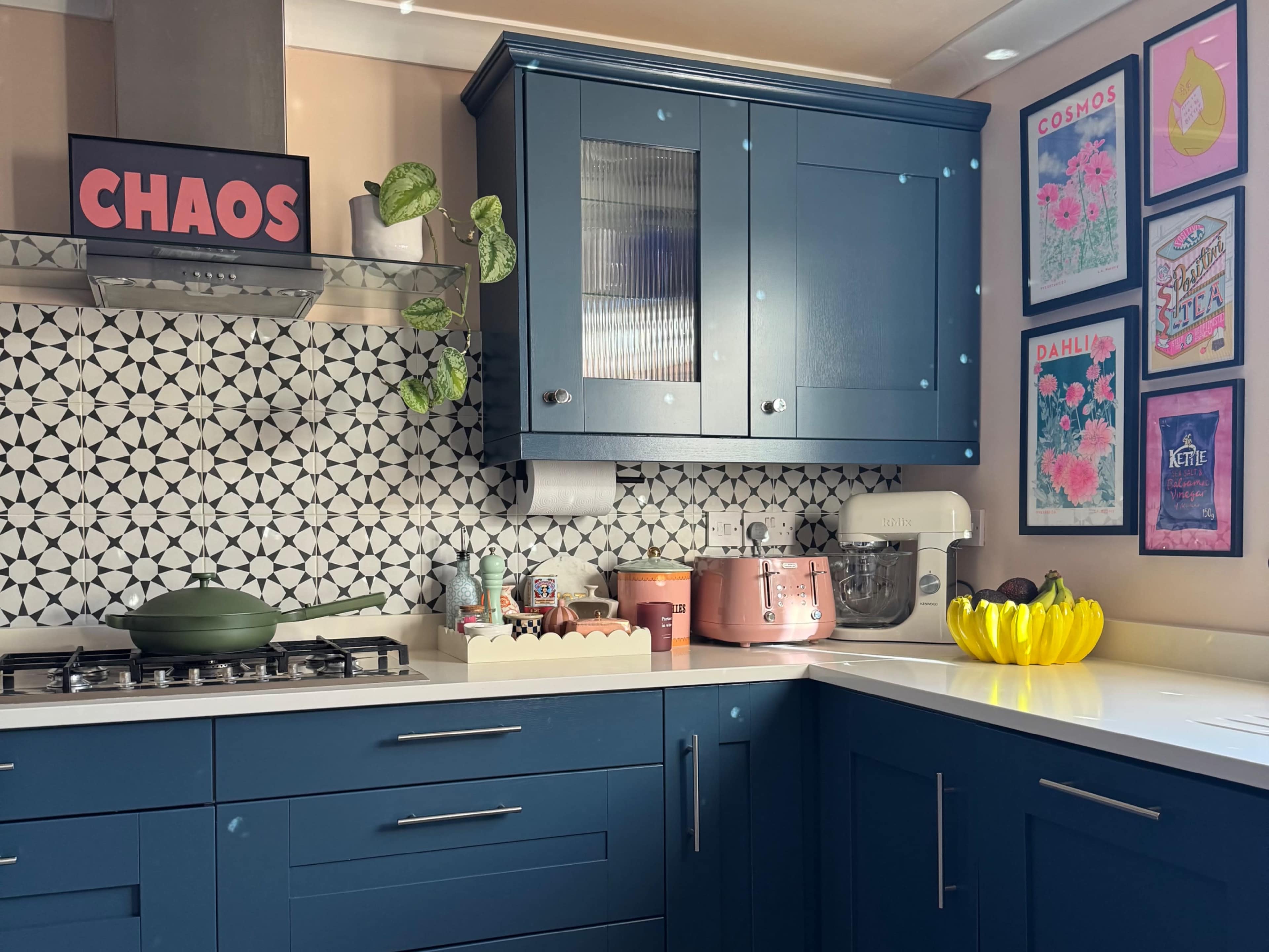 The image shows a modern kitchen with blue cabinetry, a patterned backsplash, and colorful artwork on the walls.