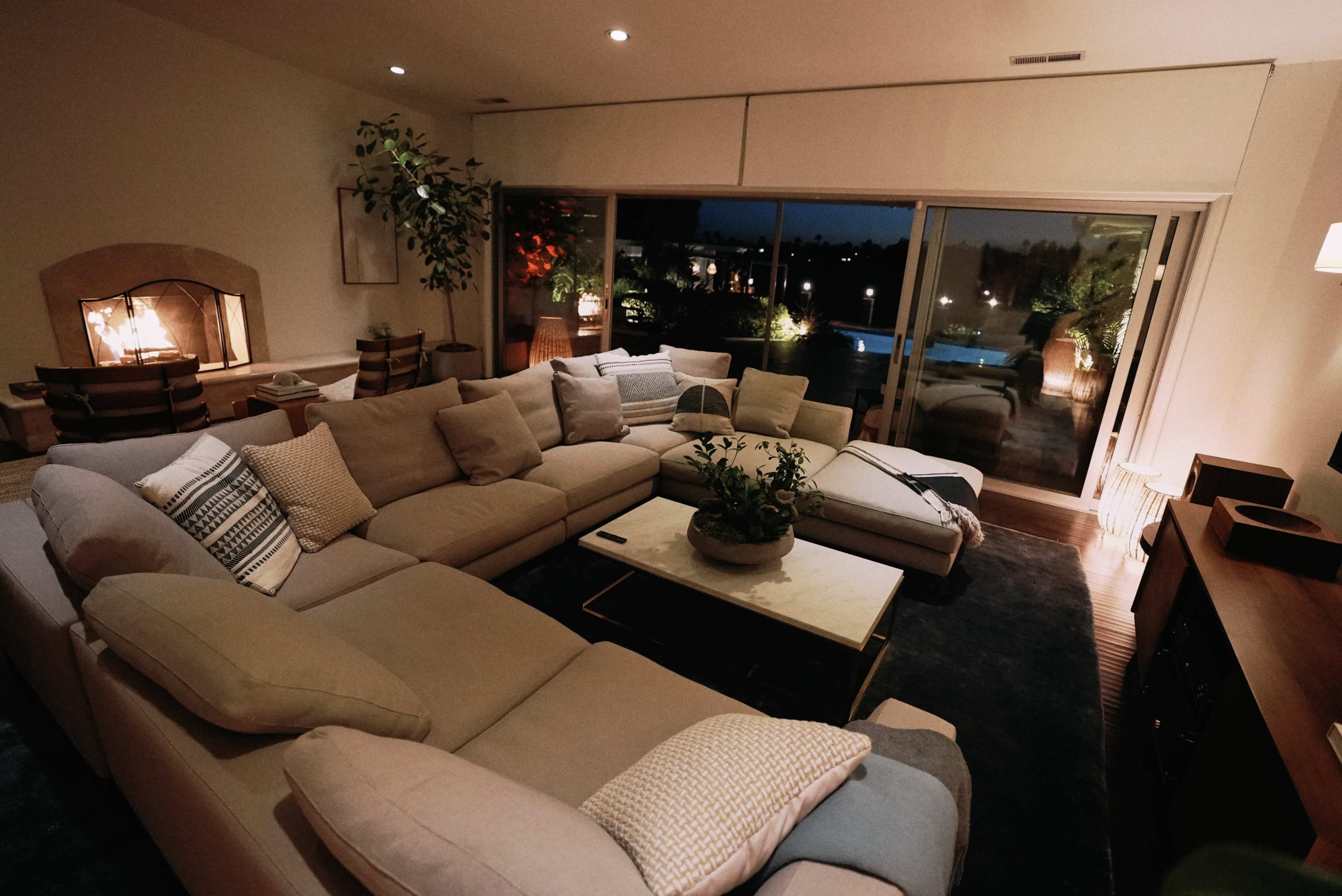 A spacious, modern living room features a large L-shaped sectional sofa, a coffee table with a plant, and sliding glass doors that open to an outdoor area with pool lighting.
