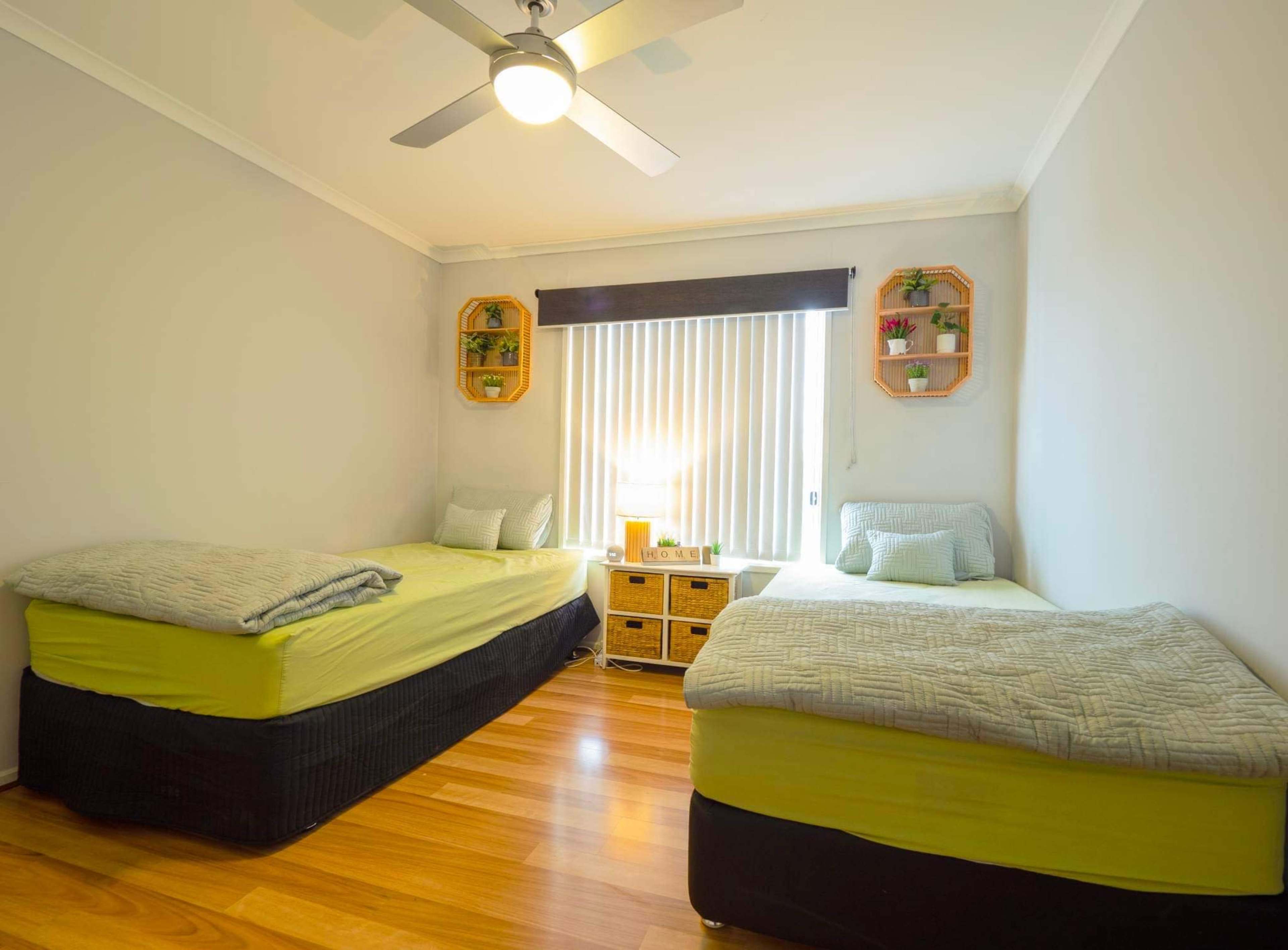 The room features two single beds with light green bedding, a window with vertical blinds, and a wooden dresser adorned with small plants and a lamp.