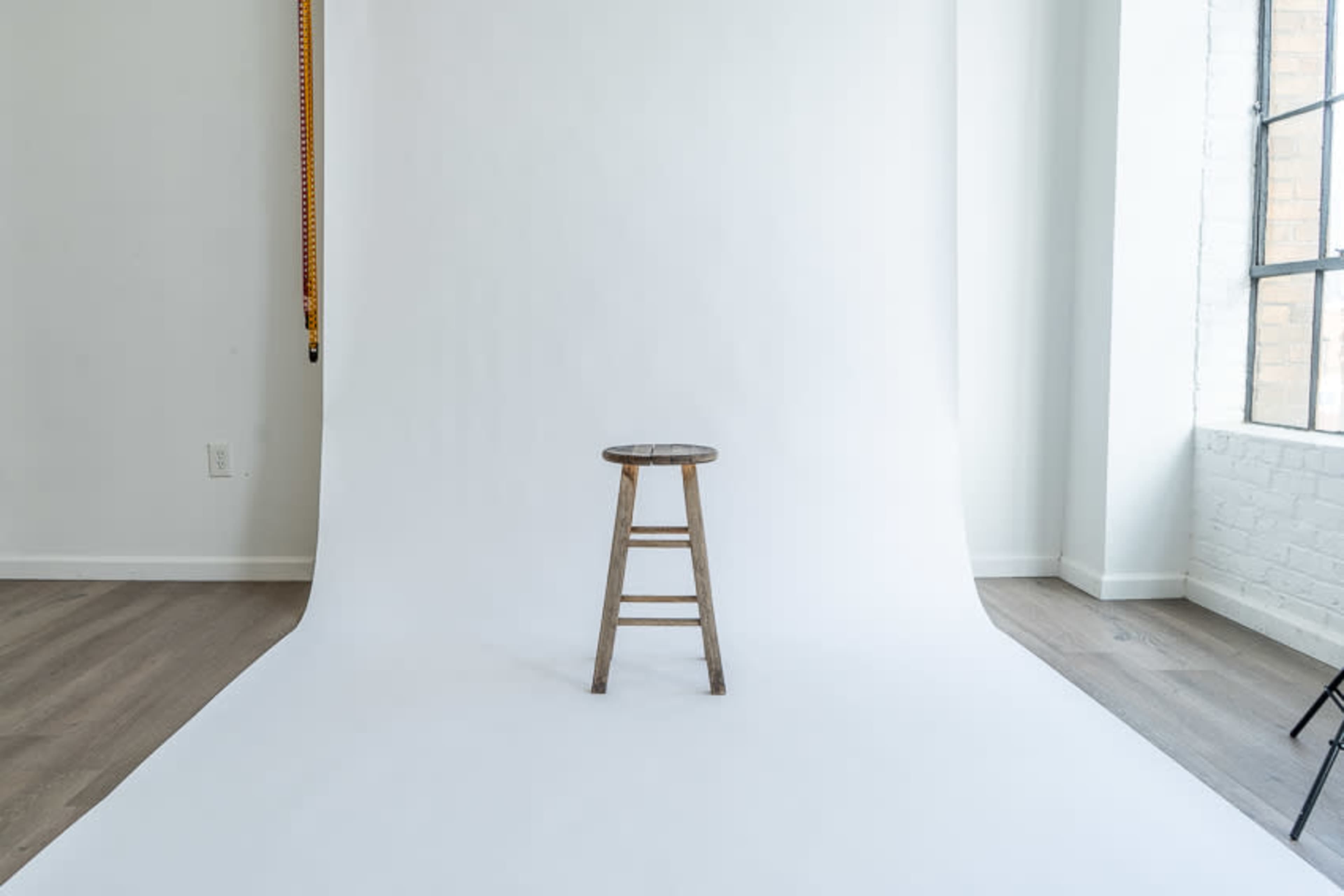 A wooden stool stands on a white backdrop in a well-lit room with large windows.