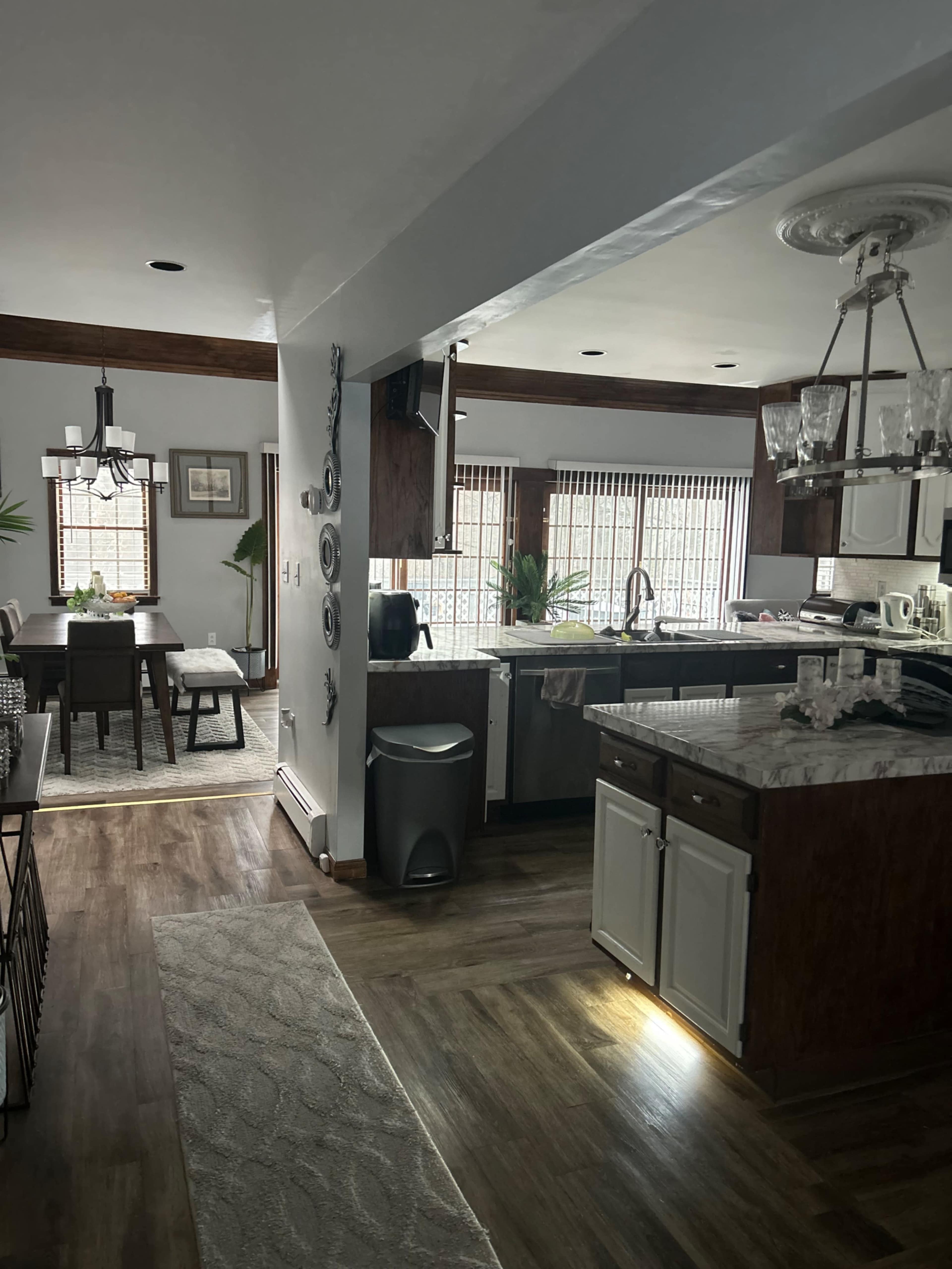 The image shows a kitchen and dining area with wooden cabinets, a dining table, and large windows with blinds.
