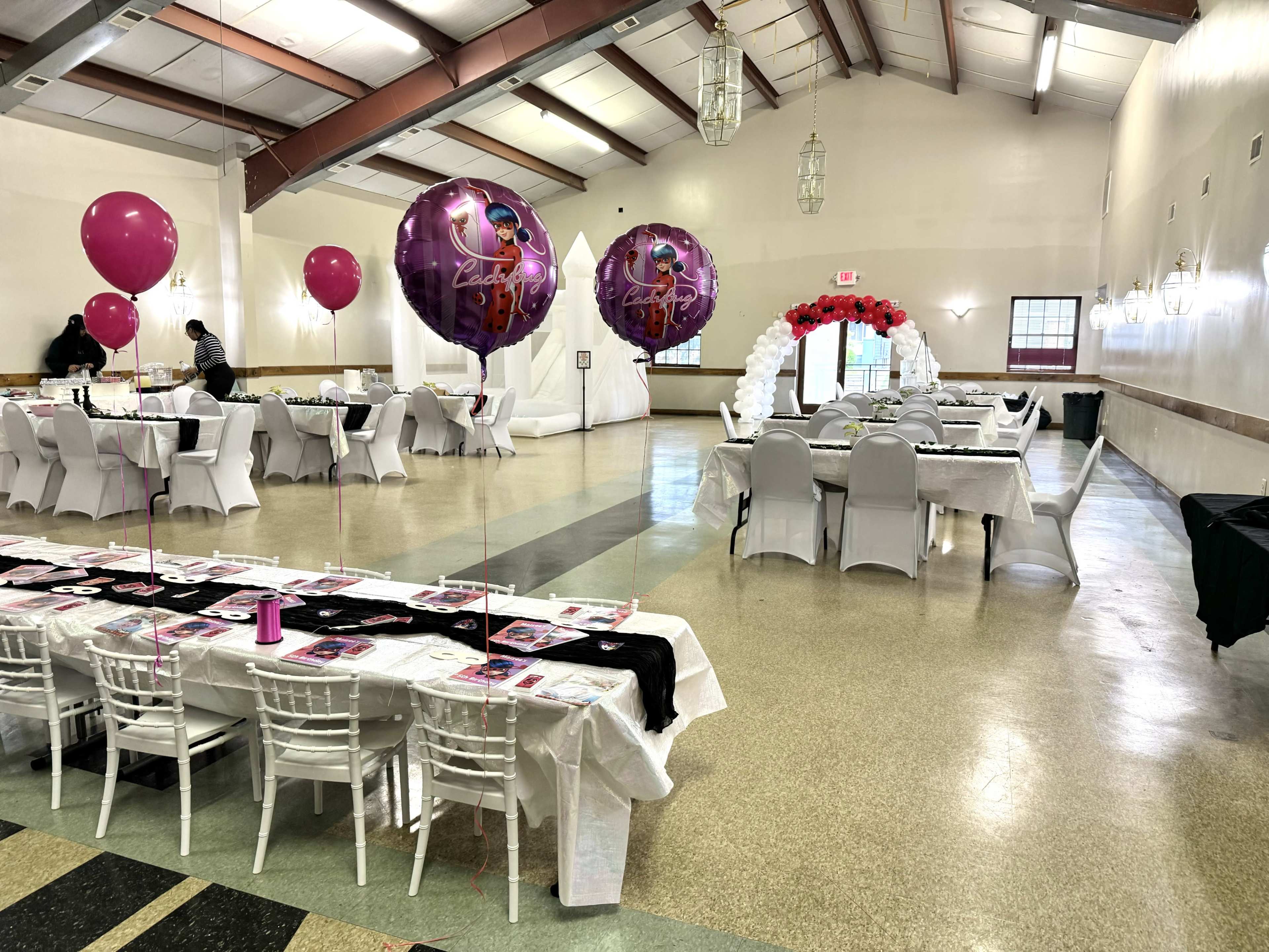 A banquet hall set up for a celebration, featuring decorated tables, large balloons, and a floral archway.