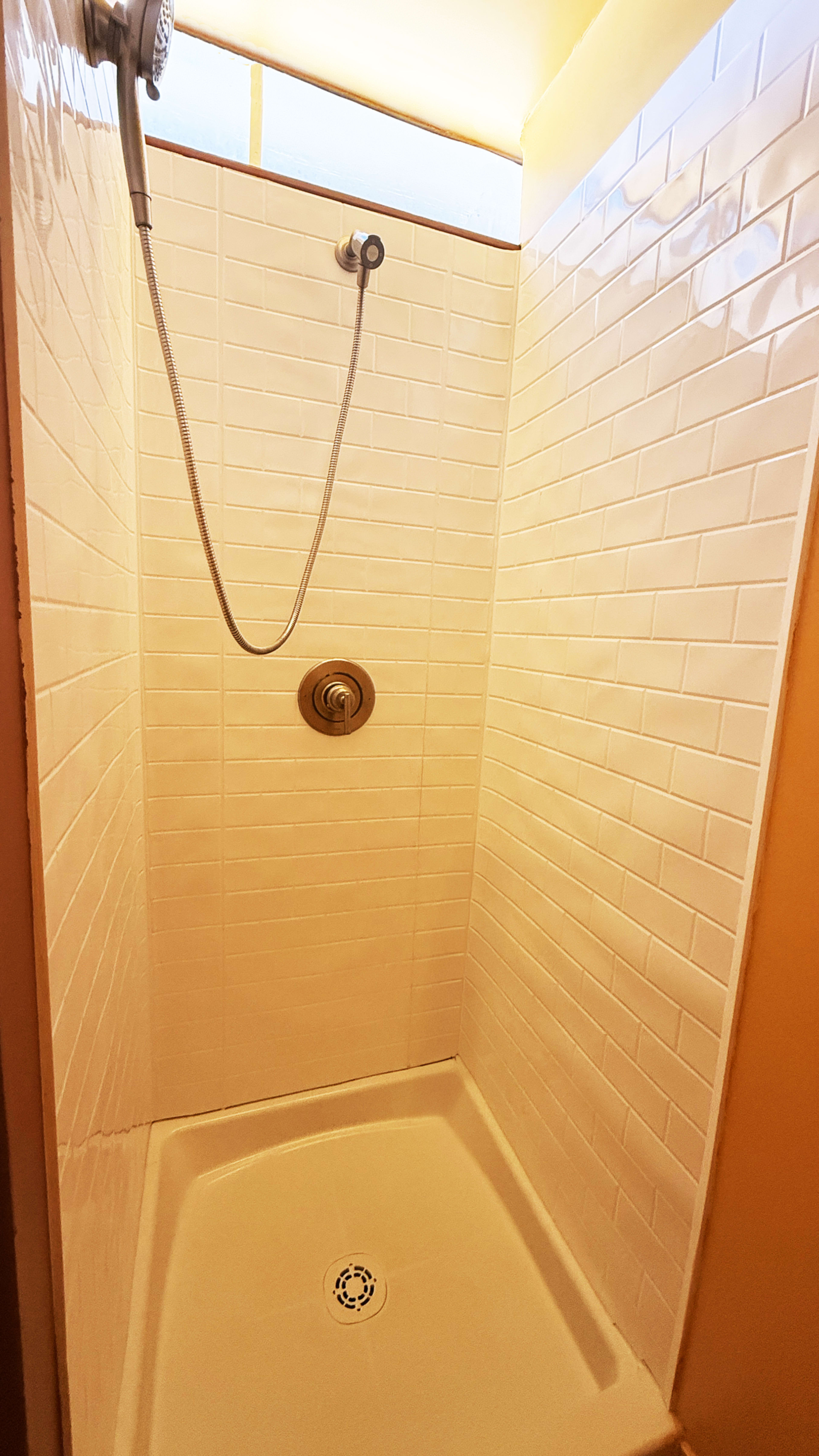 The image shows a small, tiled shower enclosure with a handheld showerhead and a drain on the shower floor.