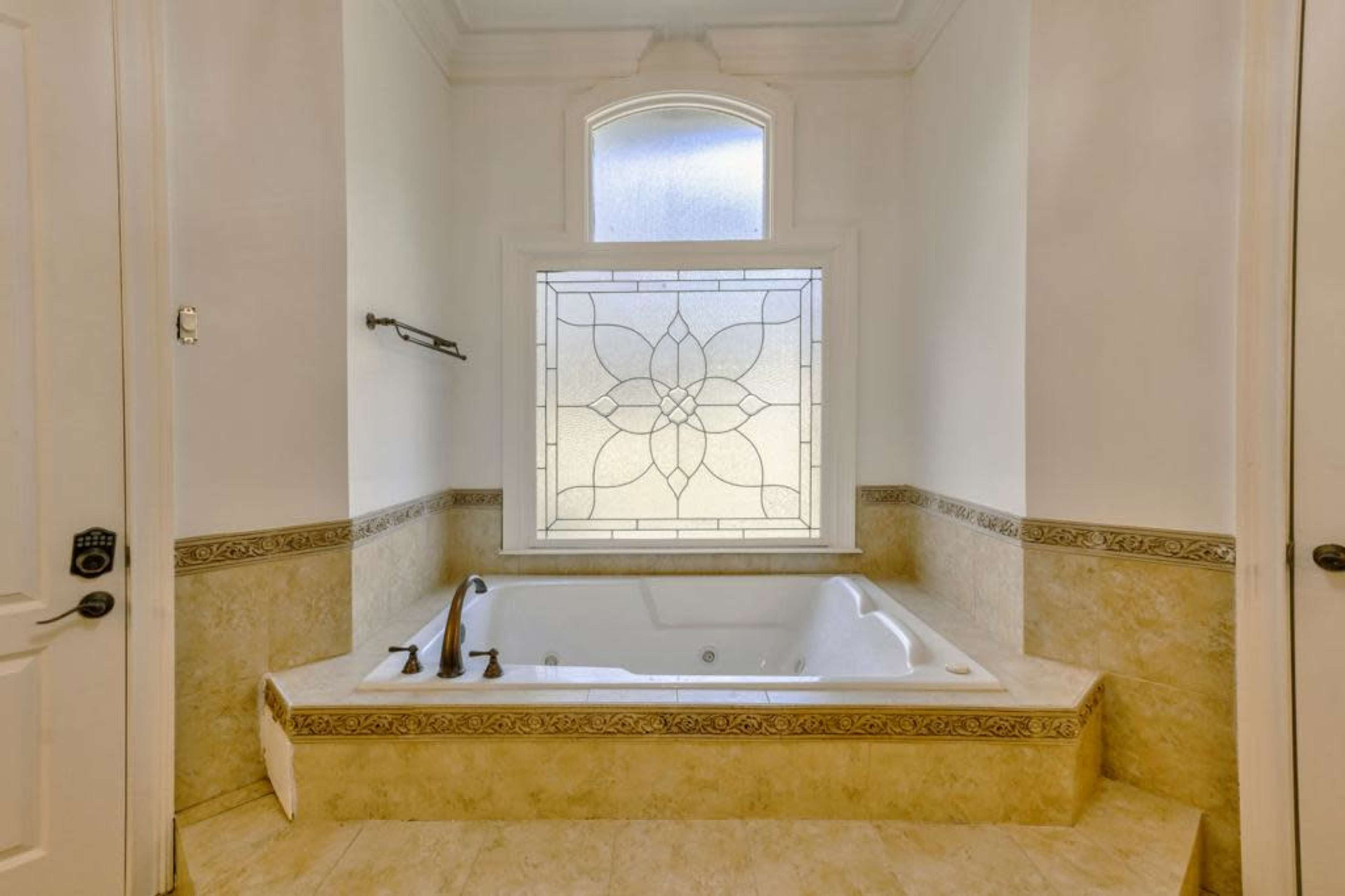 A spacious bathroom featuring a jetted tub beneath a decorative window surrounded by light-colored walls and patterned tiles.