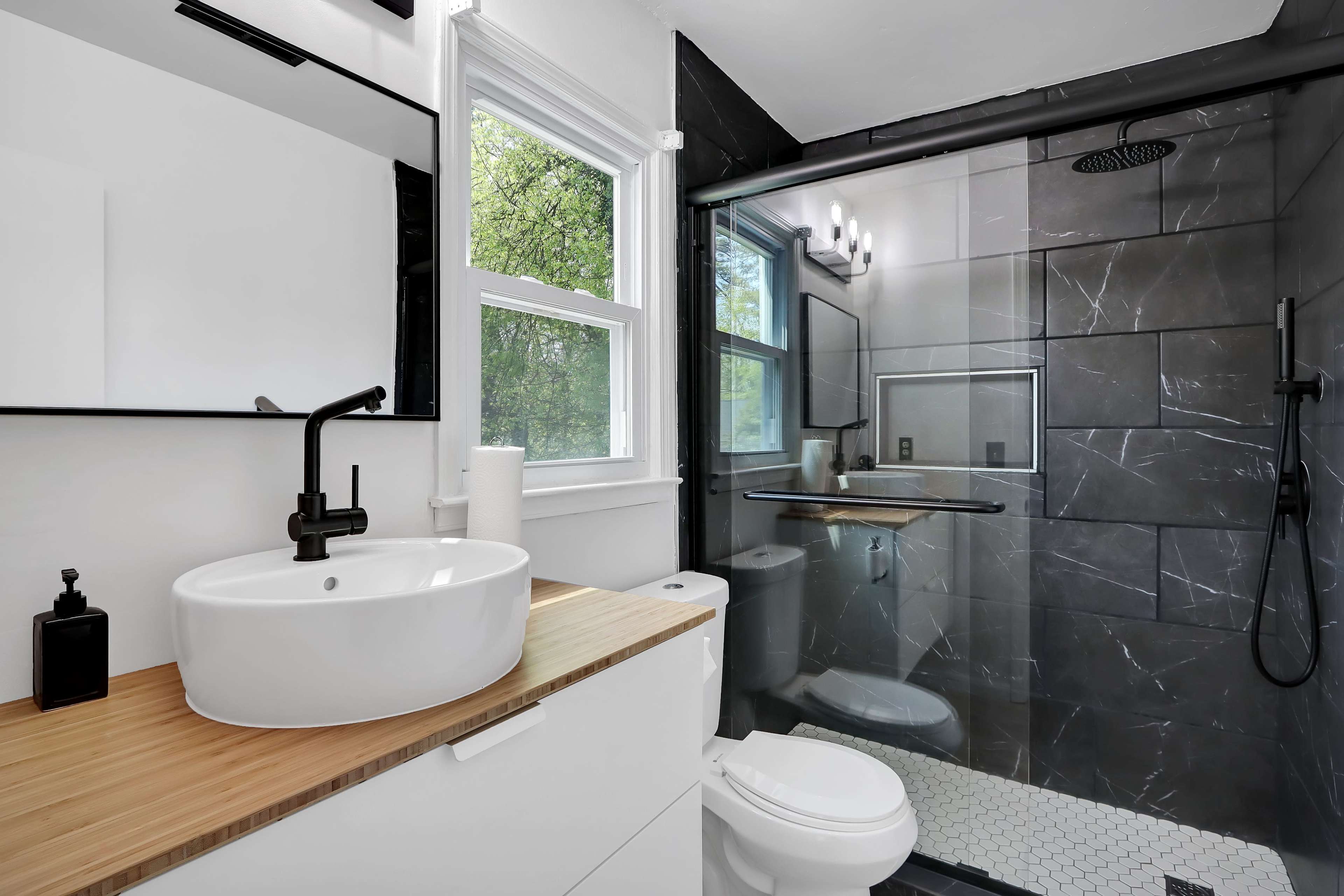 The bathroom features a modern design with a circular sink on a wooden countertop, a glass shower enclosure, and a combination of white and black tile walls.
