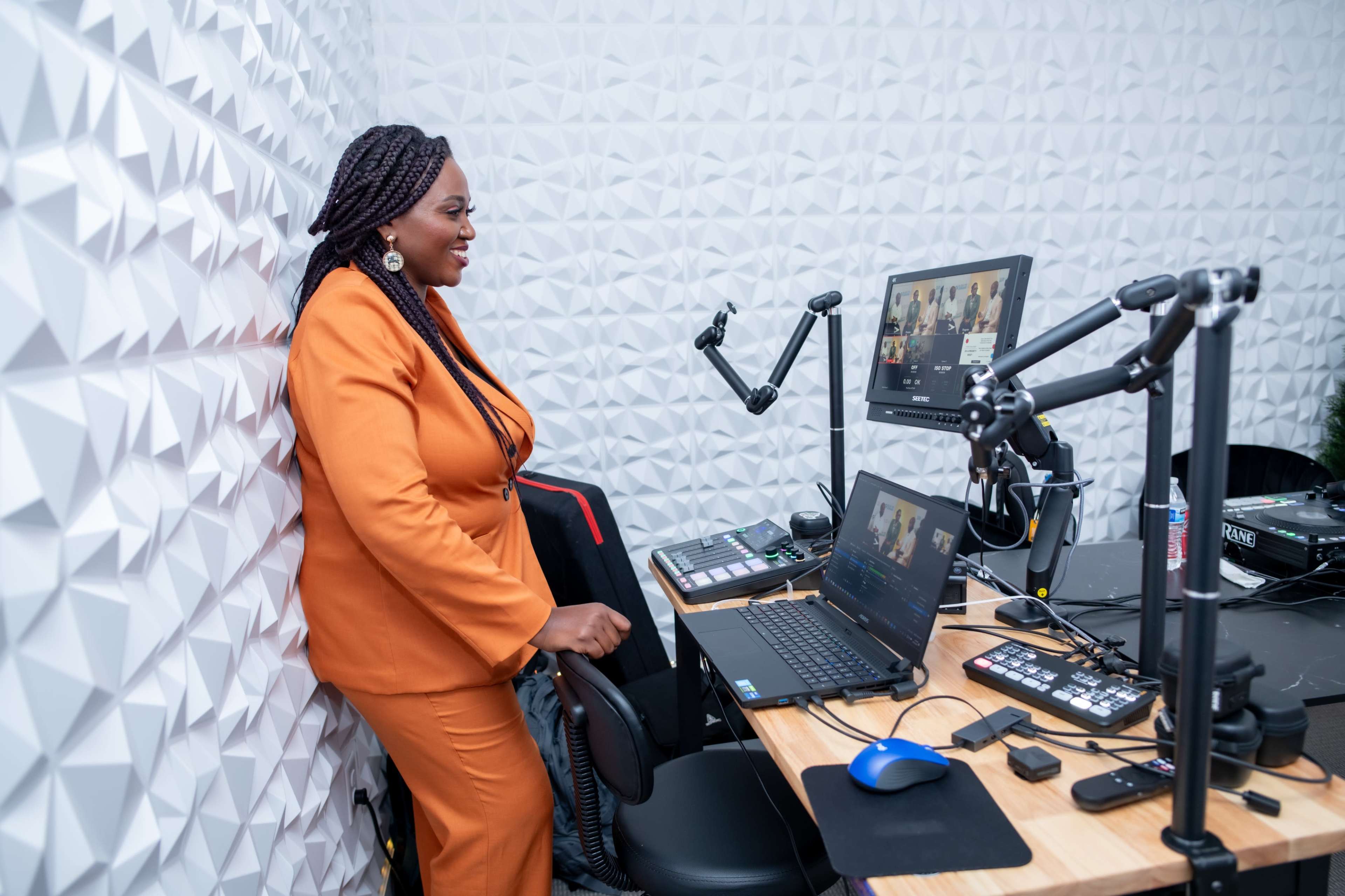 A woman in an orange suit stands beside a workstation featuring multiple monitors and audio equipment in a modern studio setting.