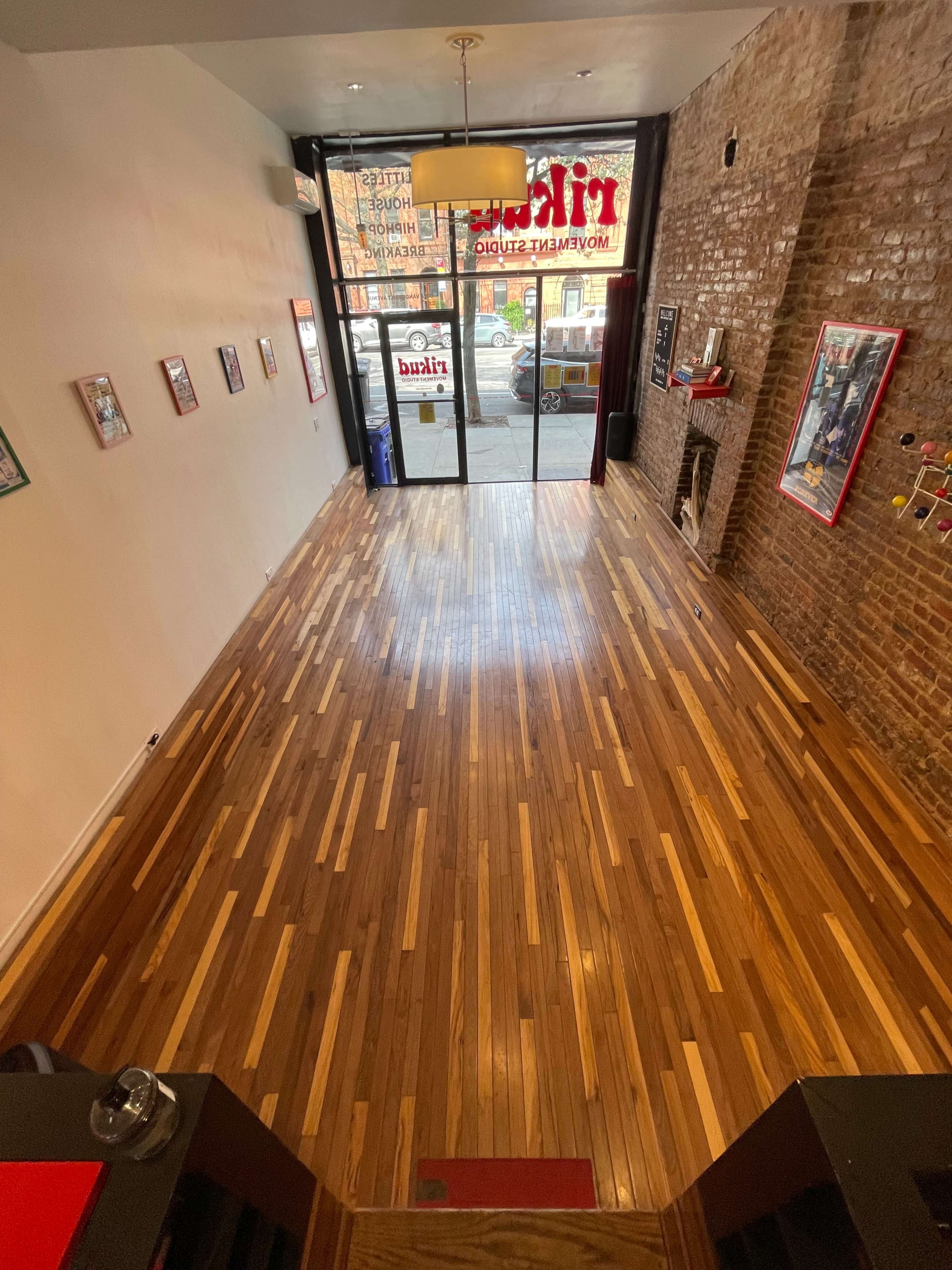 The image shows an empty interior space with a polished wooden floor, large windows at the front, and brick walls adorned with framed pictures.