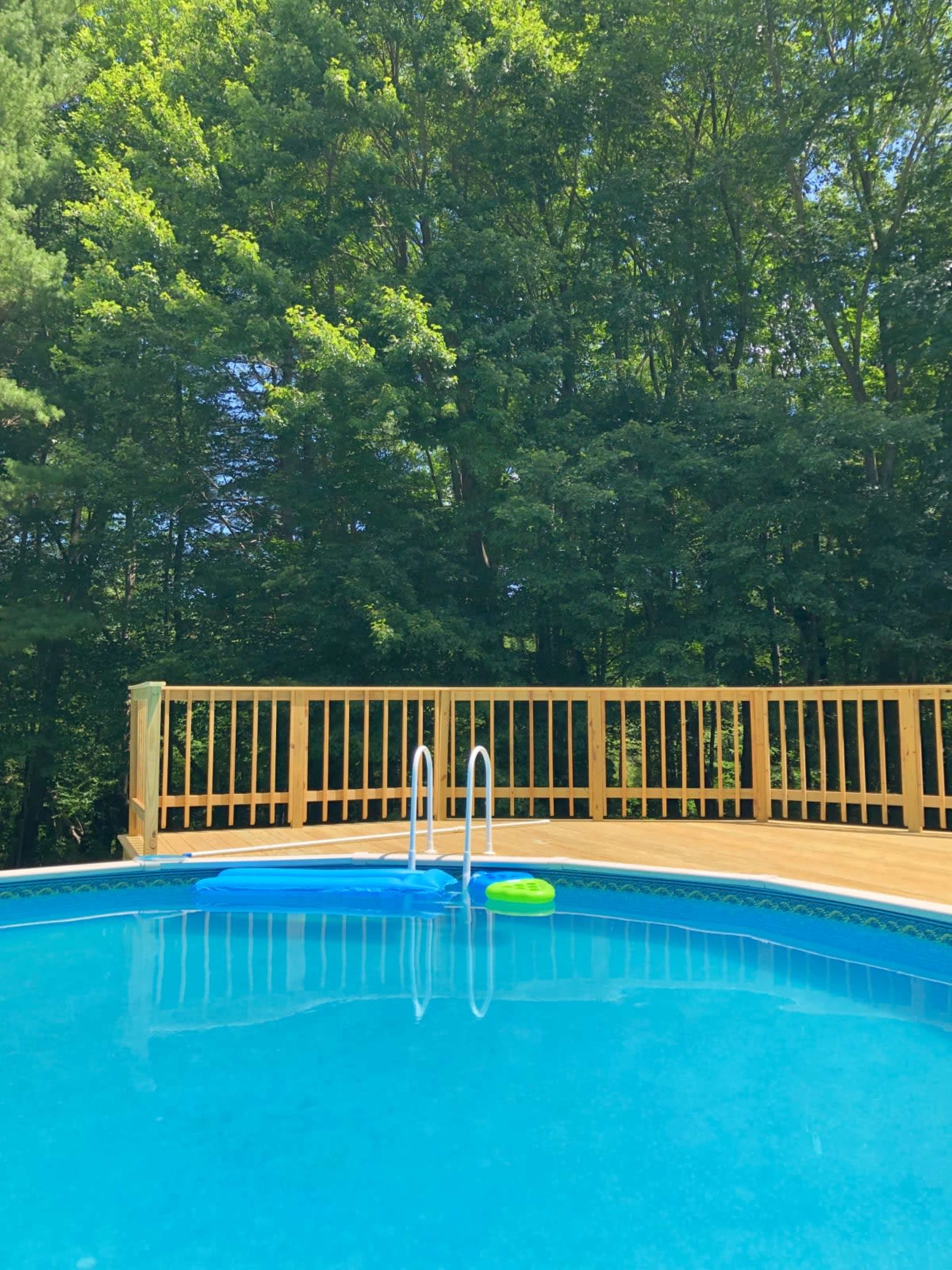 A clear swimming pool is surrounded by a wooden deck and green trees in the background.