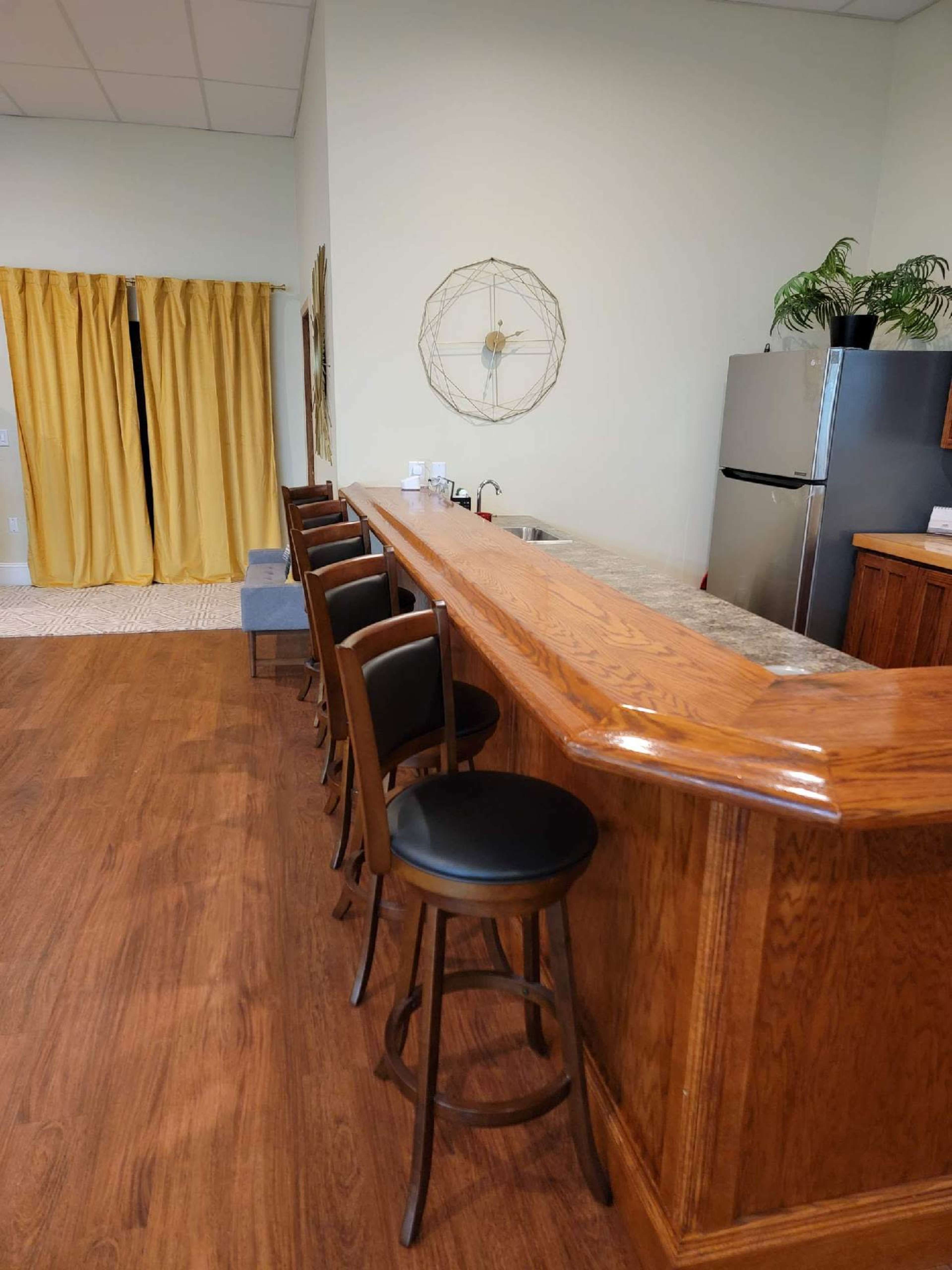 A wooden bar with high stools, set against a backdrop of yellow curtains and a modern kitchen area featuring a refrigerator.