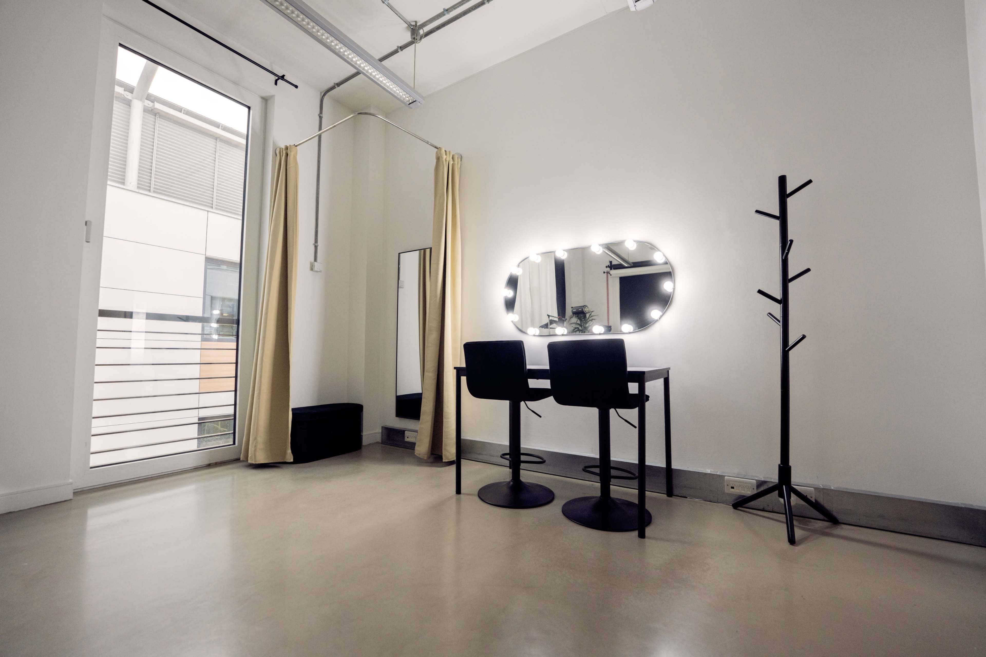 A minimalist room with a table and two chairs, a large illuminated mirror, a coat rack, and a floor-to-ceiling window framed by sheer curtains.