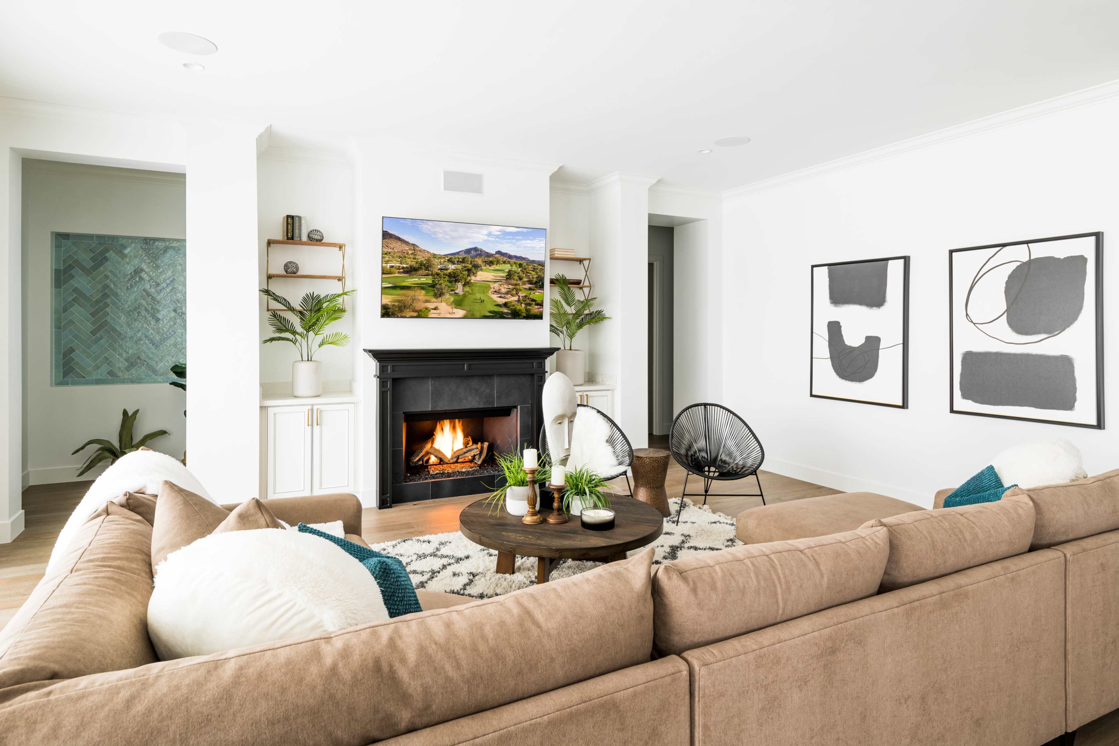 A modern living room features a brown sectional couch, a round coffee table with plants, and a fireplace with a mounted television above it, surrounded by decorative artwork and houseplants.