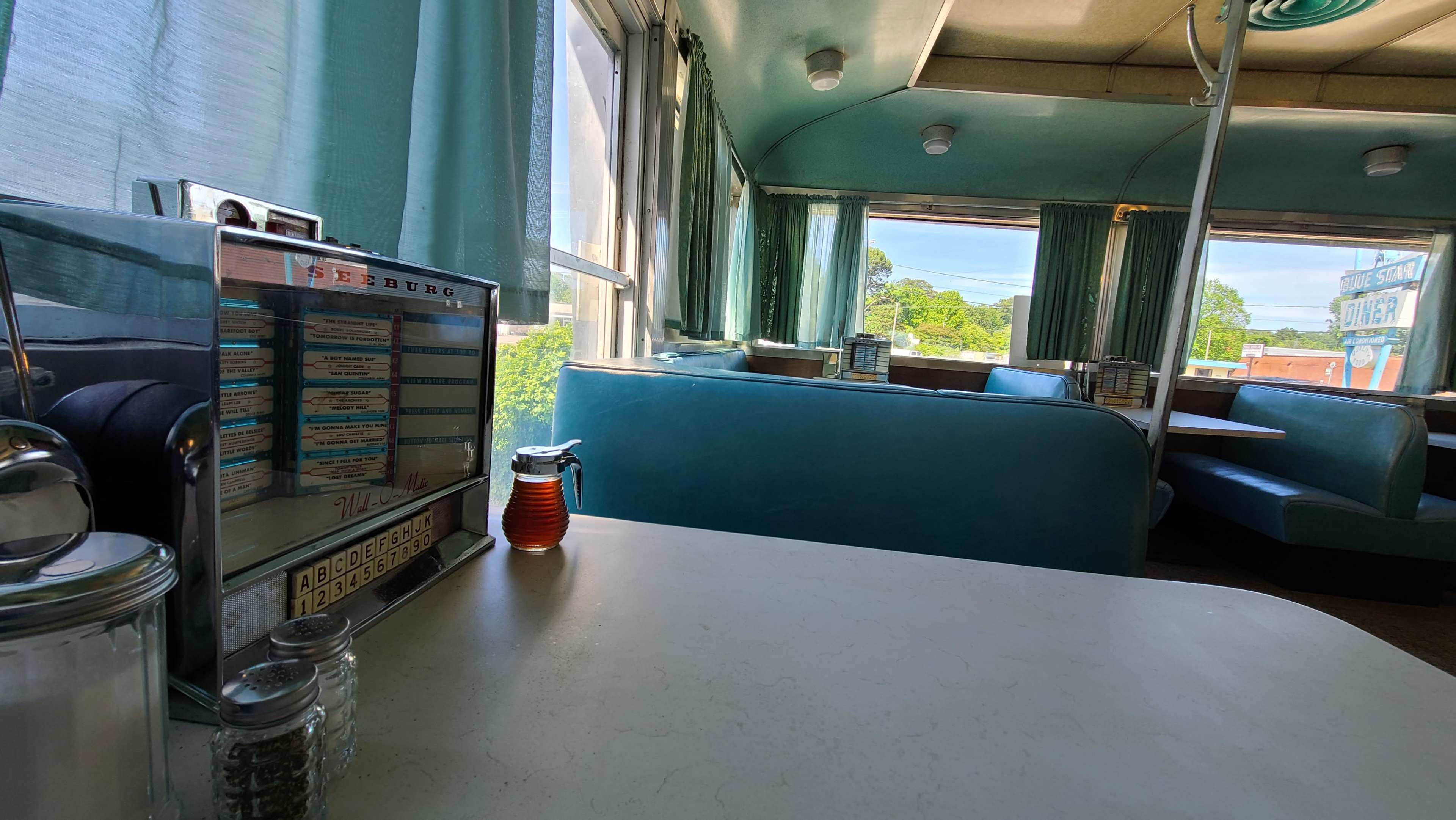The interior of a diner with teal booths, large windows draped in light green curtains, and a jukebox visible in the foreground.