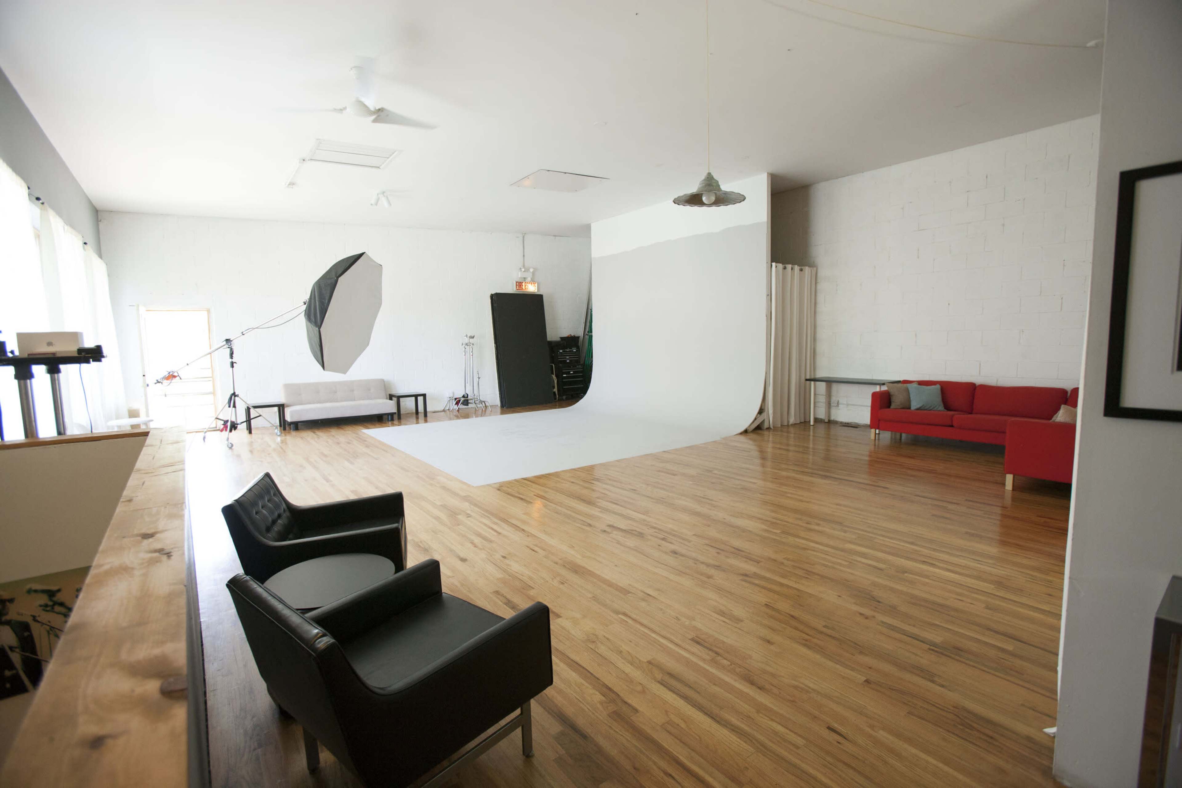 A spacious photography studio featuring a white backdrop, hardwood flooring, a red couch, and two black chairs.