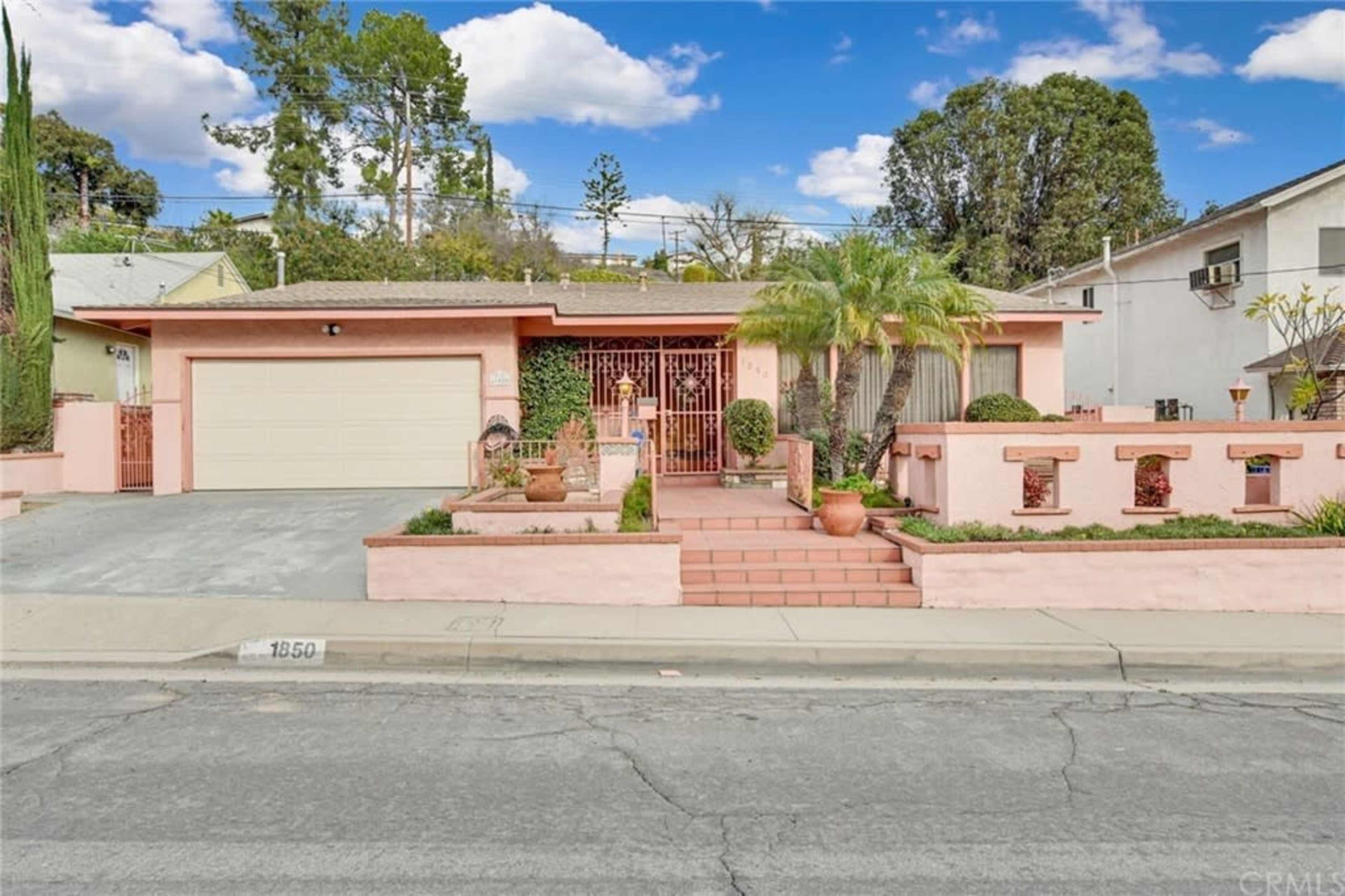 The image shows a single-story house with a pink exterior, a landscaped front yard, and a driveway leading to a garage.