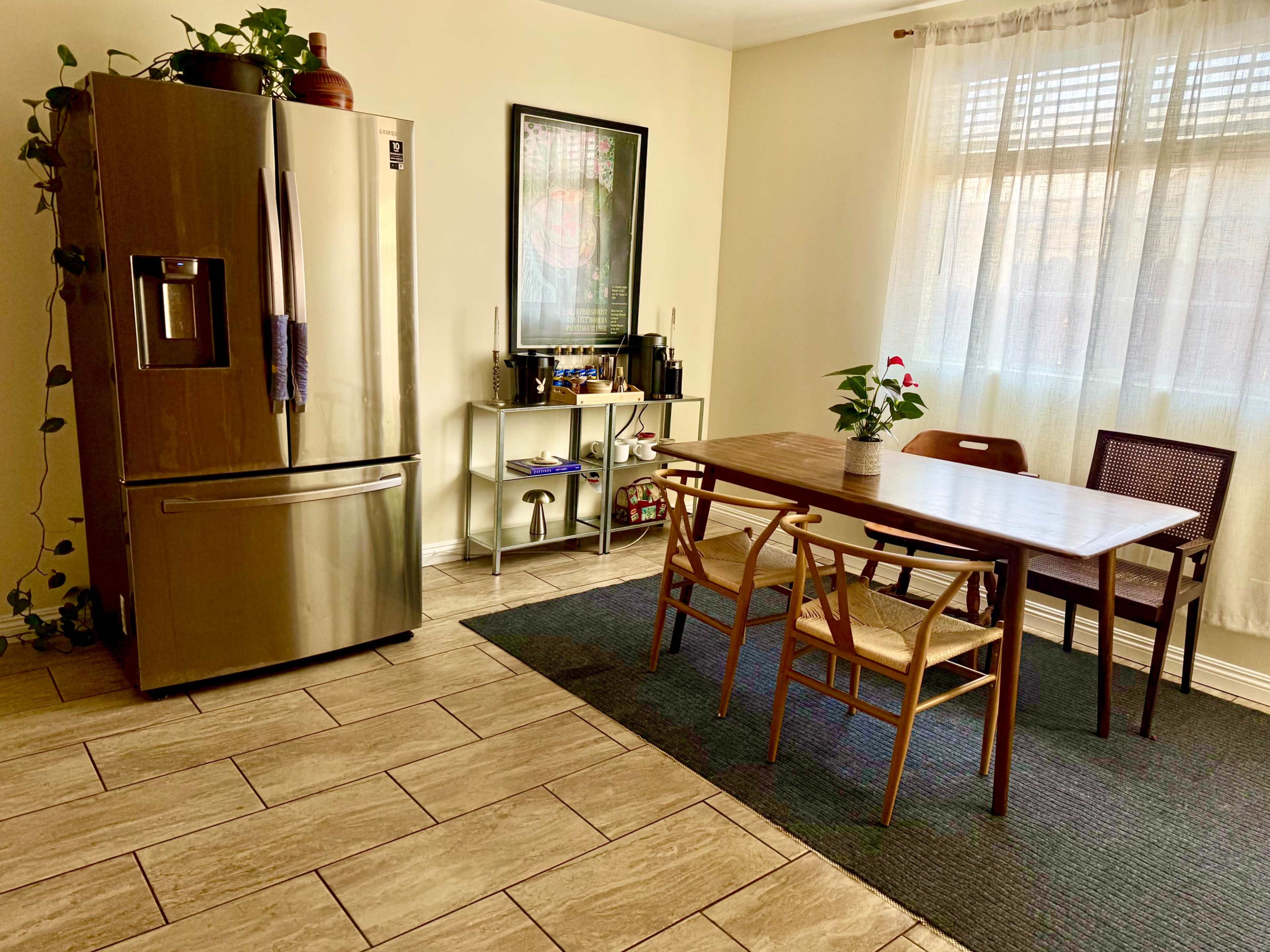 The image shows a kitchen area featuring a stainless steel refrigerator, a wooden dining table with four chairs, and a small shelf with various items.