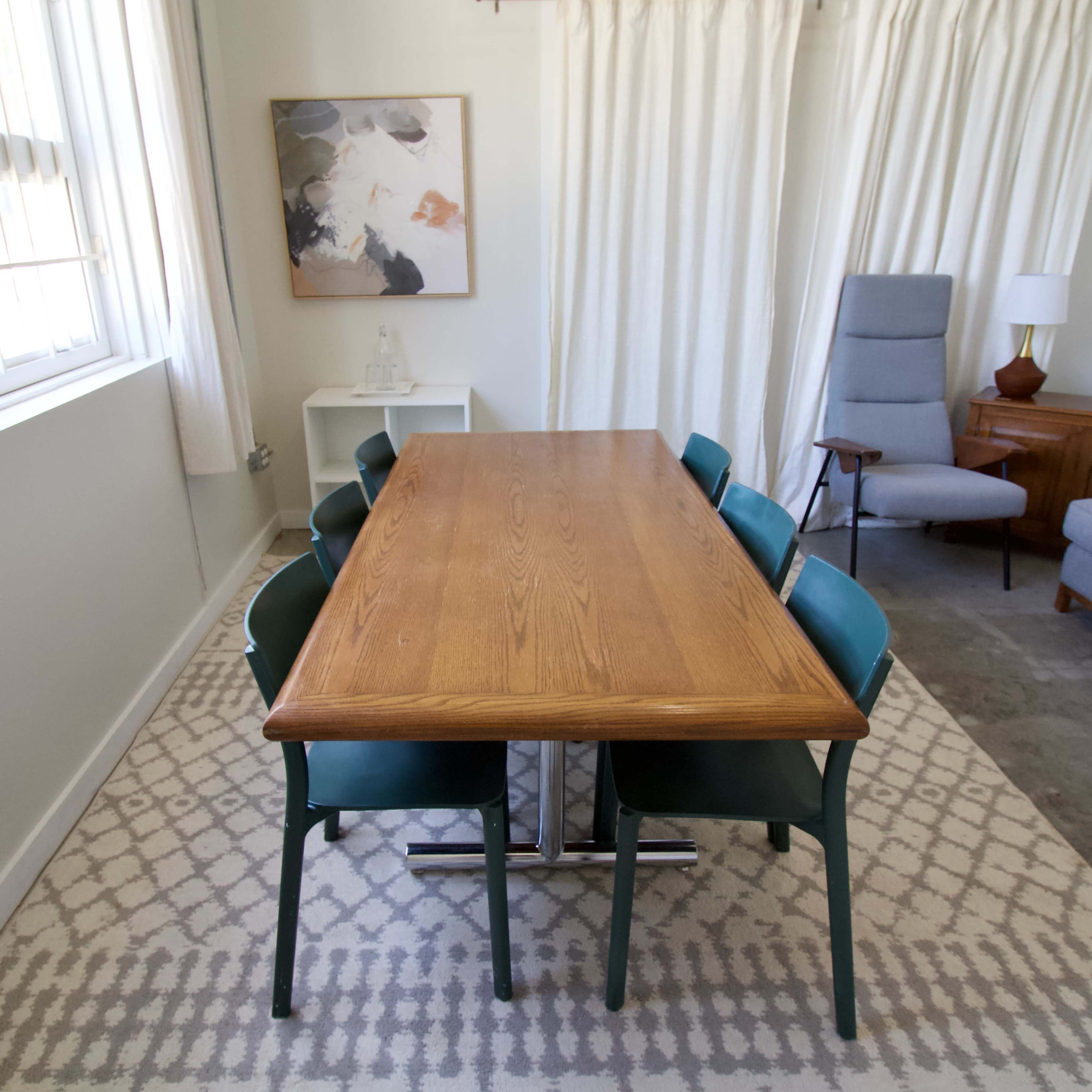 A rectangular wooden dining table with six chairs is set in a bright room featuring light-colored curtains, a painting on the wall, and an armchair in the corner.