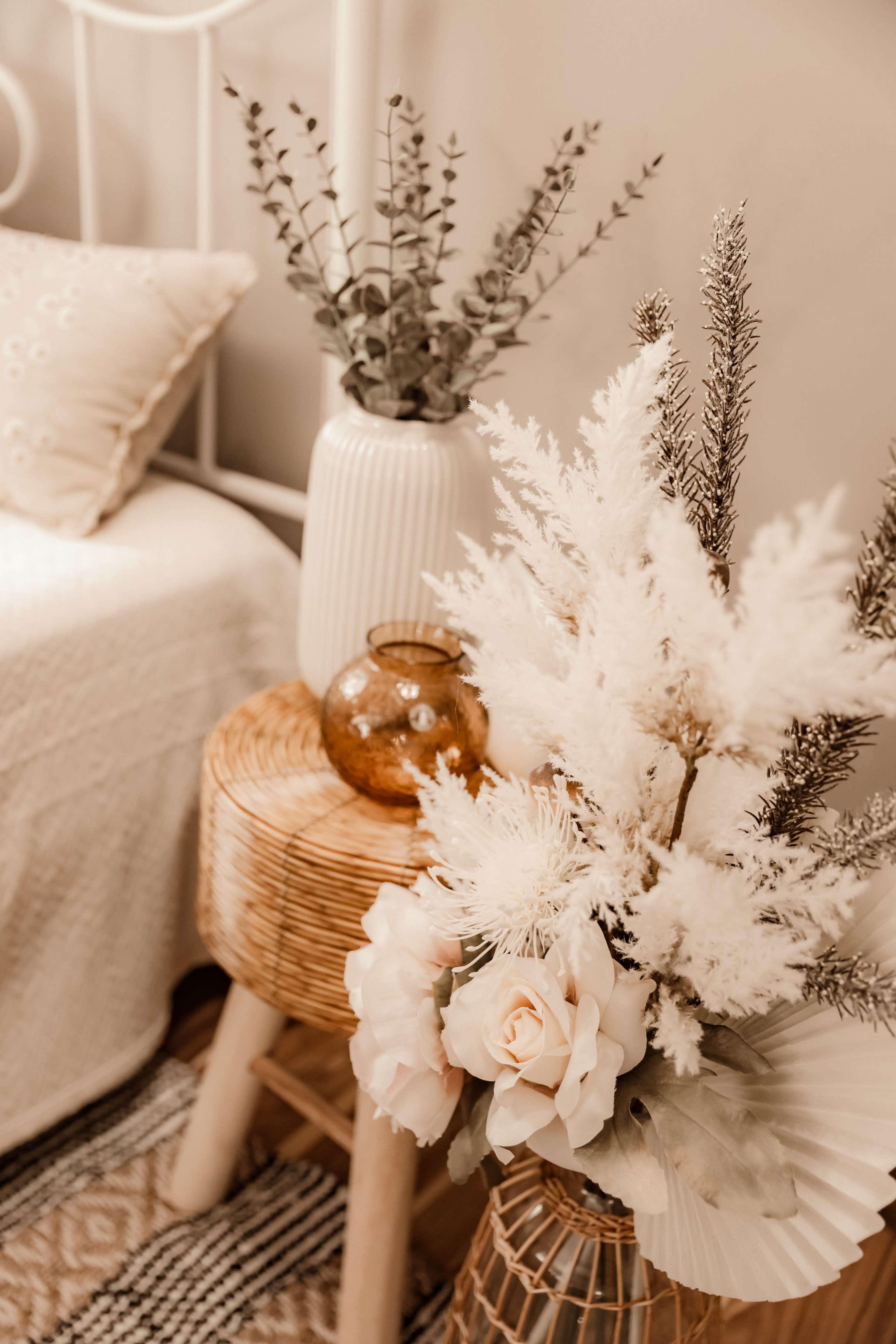 A bedside arrangement features a white floral bouquet, a brown glass vase, and two decorative stools beside a bed with a textured blanket.