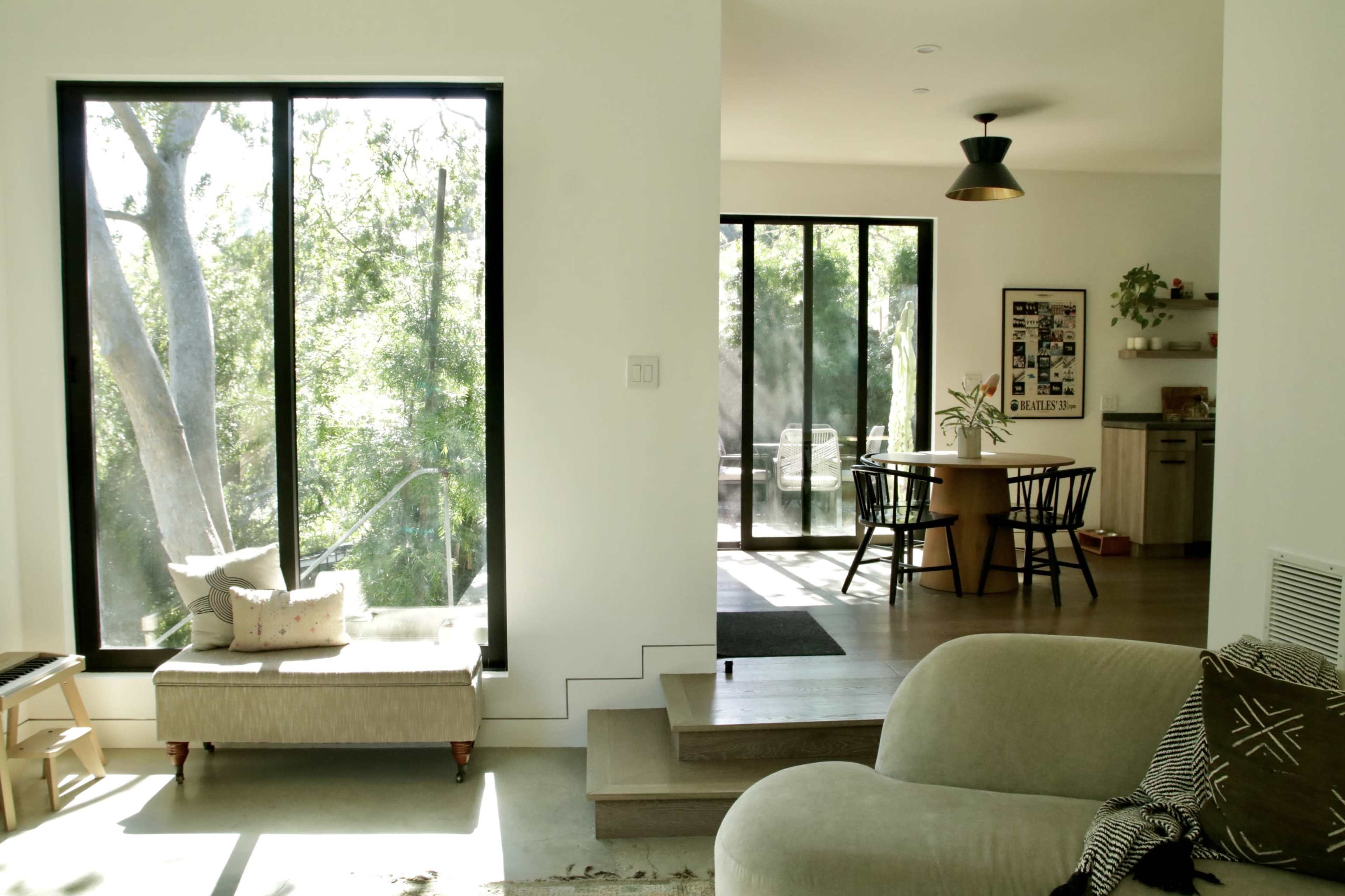The image features a modern living space with large windows allowing natural light to illuminate the room, furniture arranged for comfort, and a dining area visible in the background.