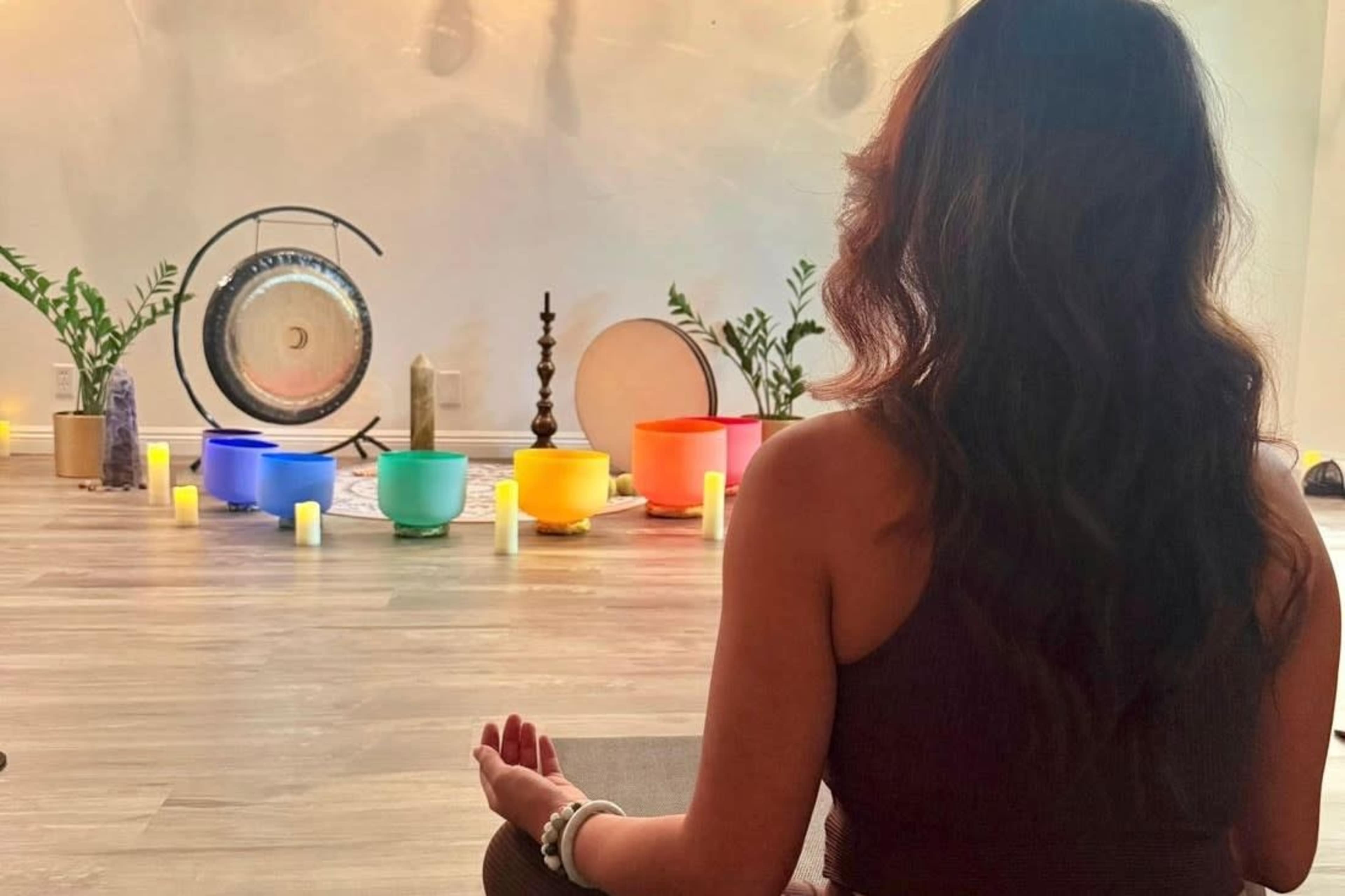 A woman sits cross-legged, facing a colorful array of sound bowls and a gong in a meditation space surrounded by greenery and candles.