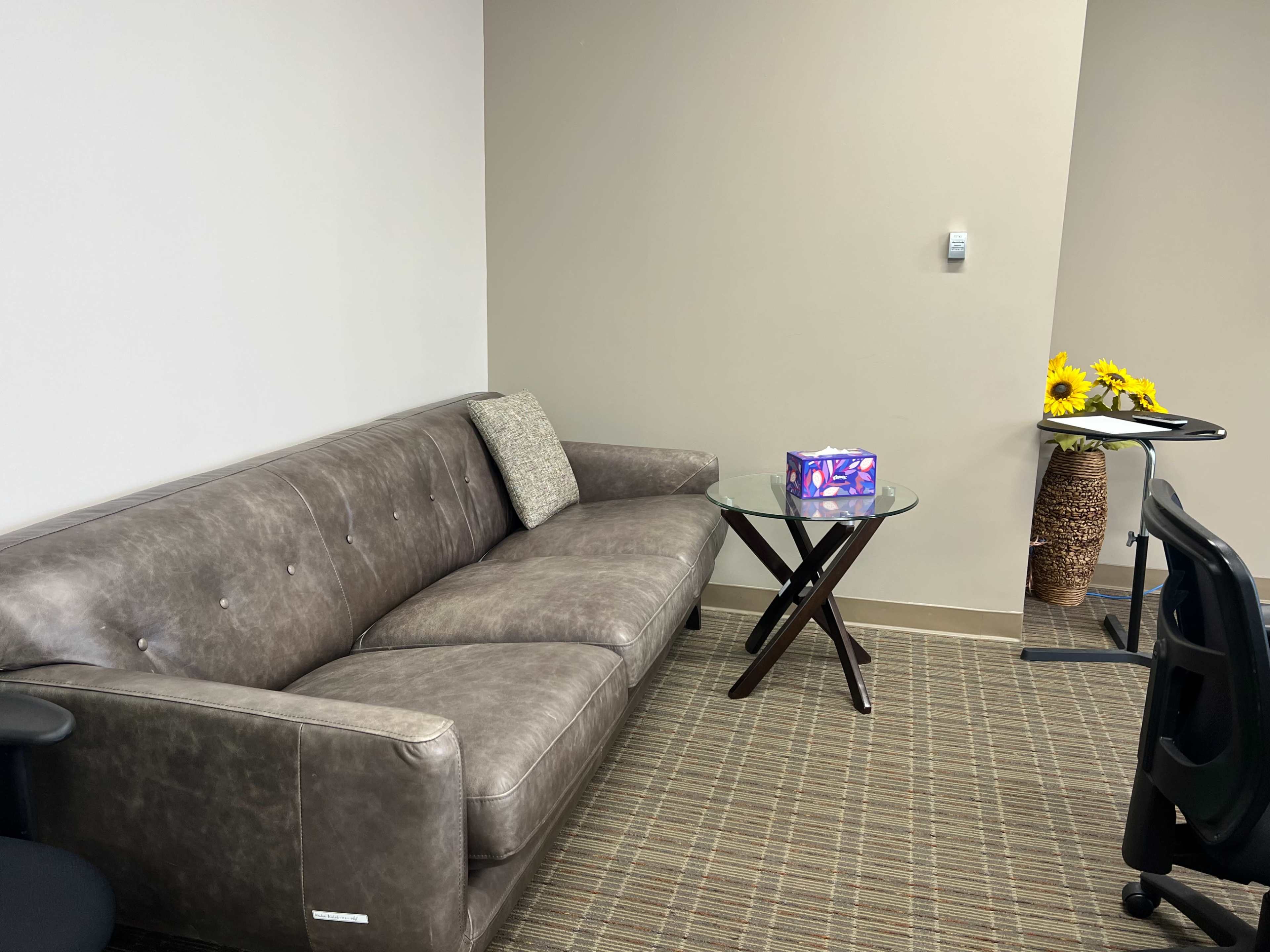 A gray couch sits beside a small glass table with a decorative box on it, while a chair is positioned nearby and a vase of sunflowers is displayed against the wall.