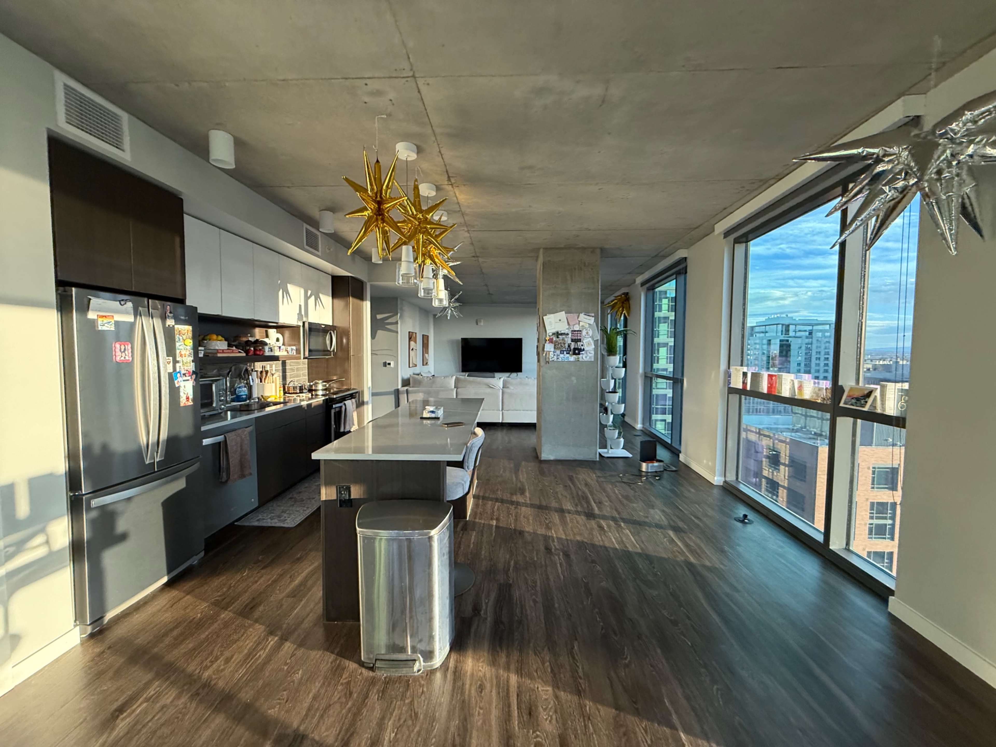 The image shows a modern, open-concept kitchen and living area with large windows and metallic star-shaped light fixtures.
