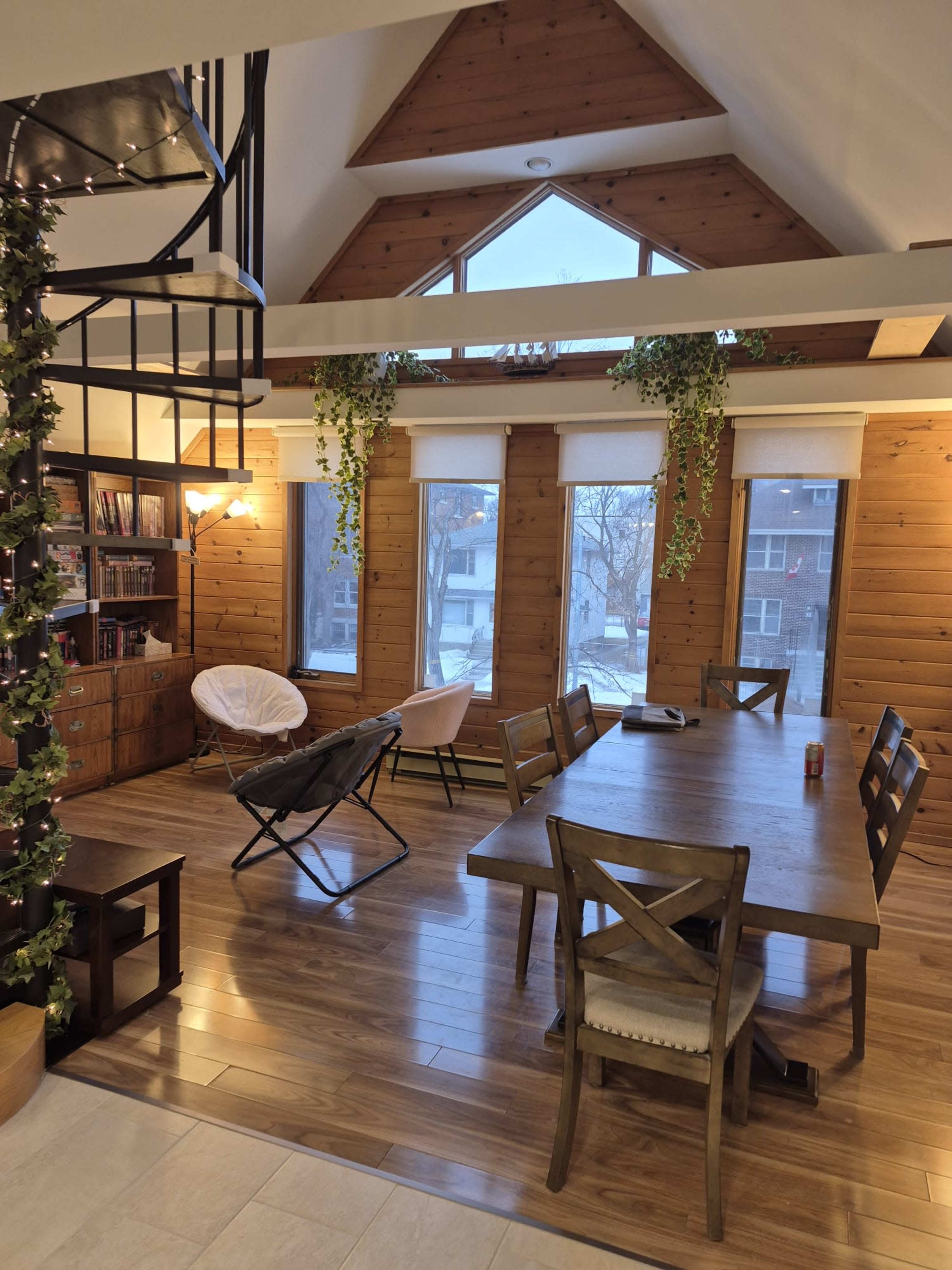 The image shows a spacious dining area with a large wooden table, surrounded by windows displaying a view of the outside, and a spiral staircase leading to an upper level.